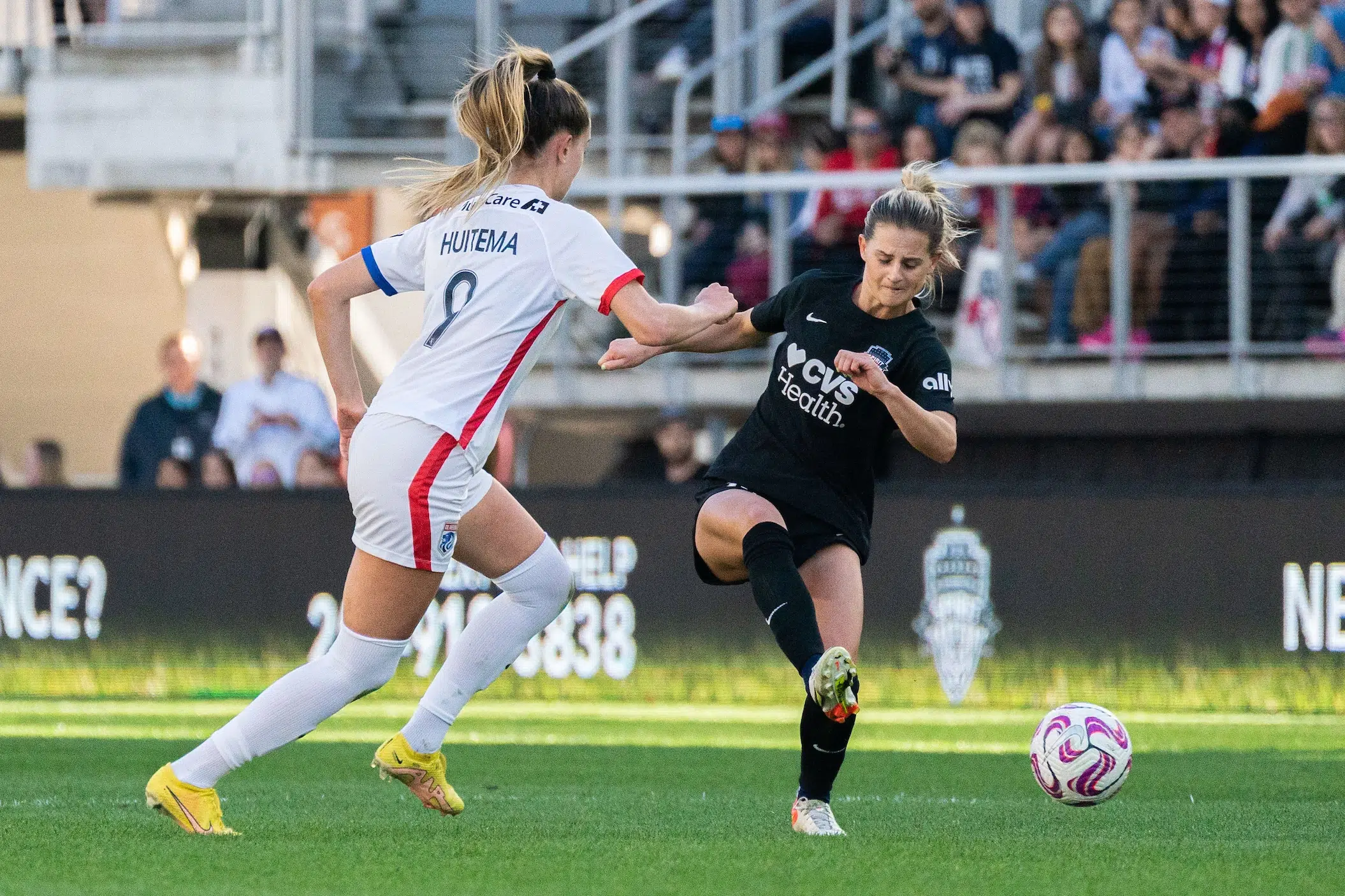 Bayley Feist in a black jersey, black shorts and black socks kicks a soccer ball past a defender in a white uniform.