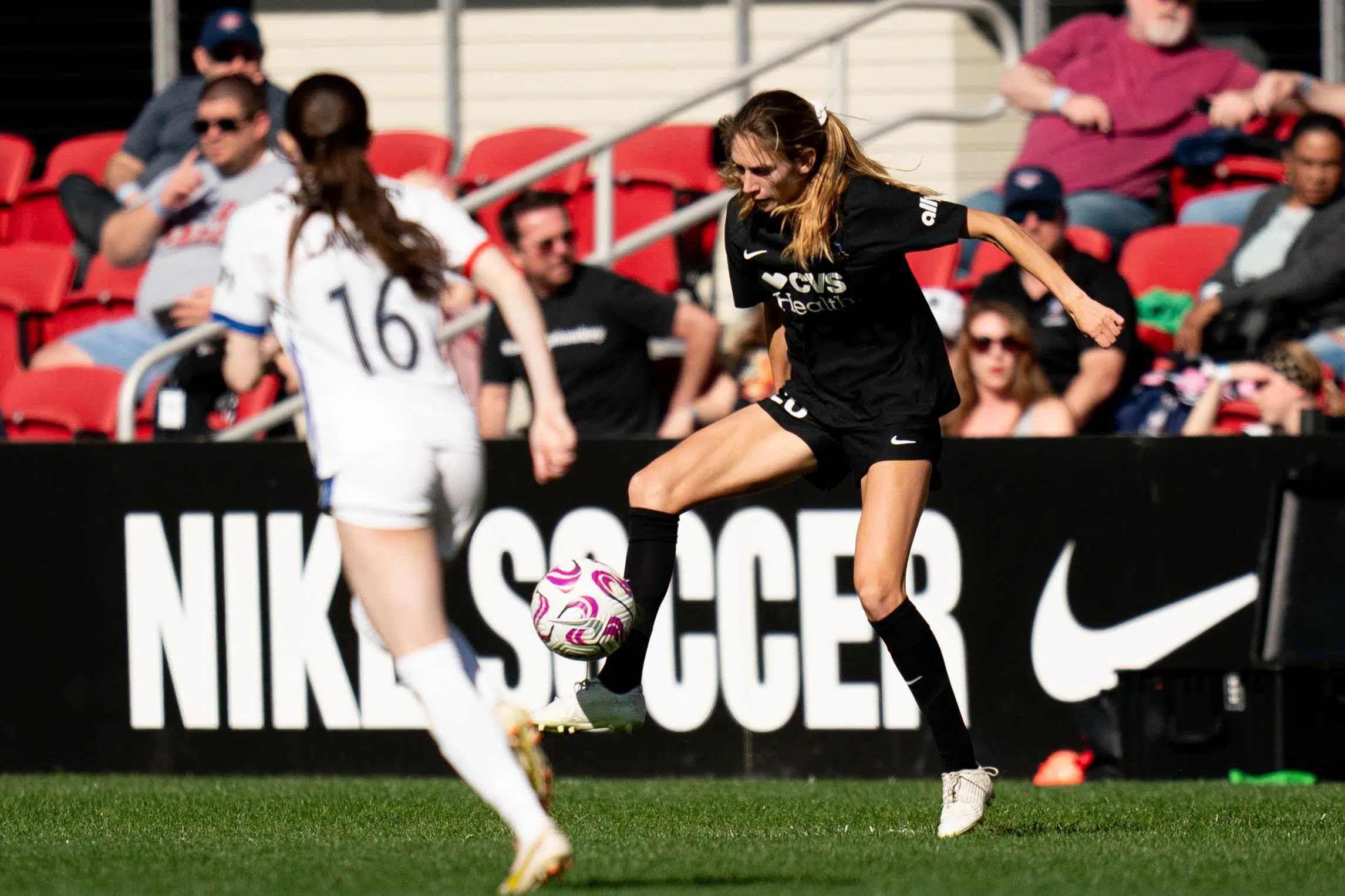 Paige Metayer in a black jersey, black shorts, black socks, and white cleats traps a soccer ball. In the background, fans watch from red seats.
