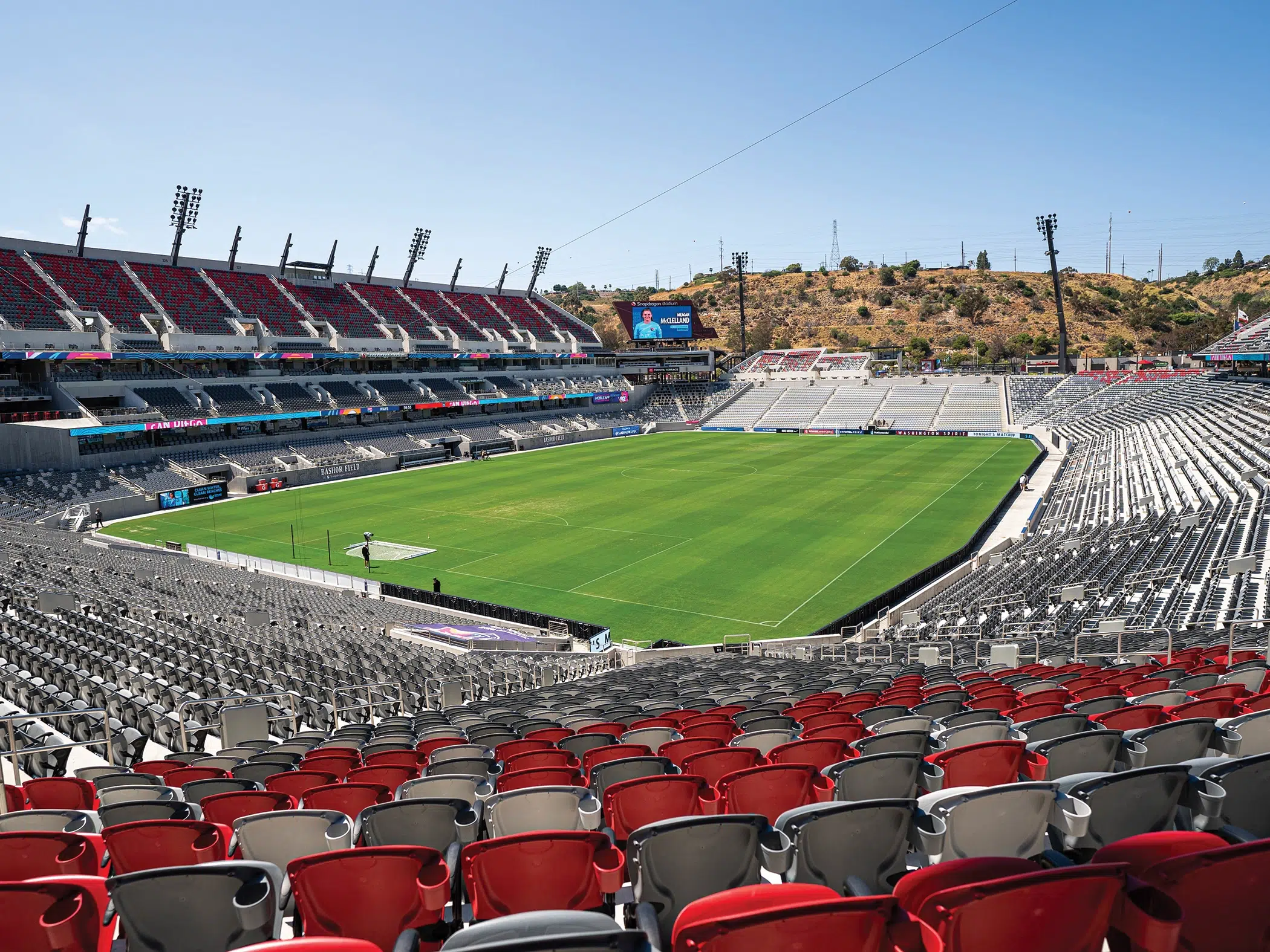 A panoramic shot of Snapdragon Stadium in San Diego. THe seats are red and gray. Rolling brown hills are seen behind the stadium.