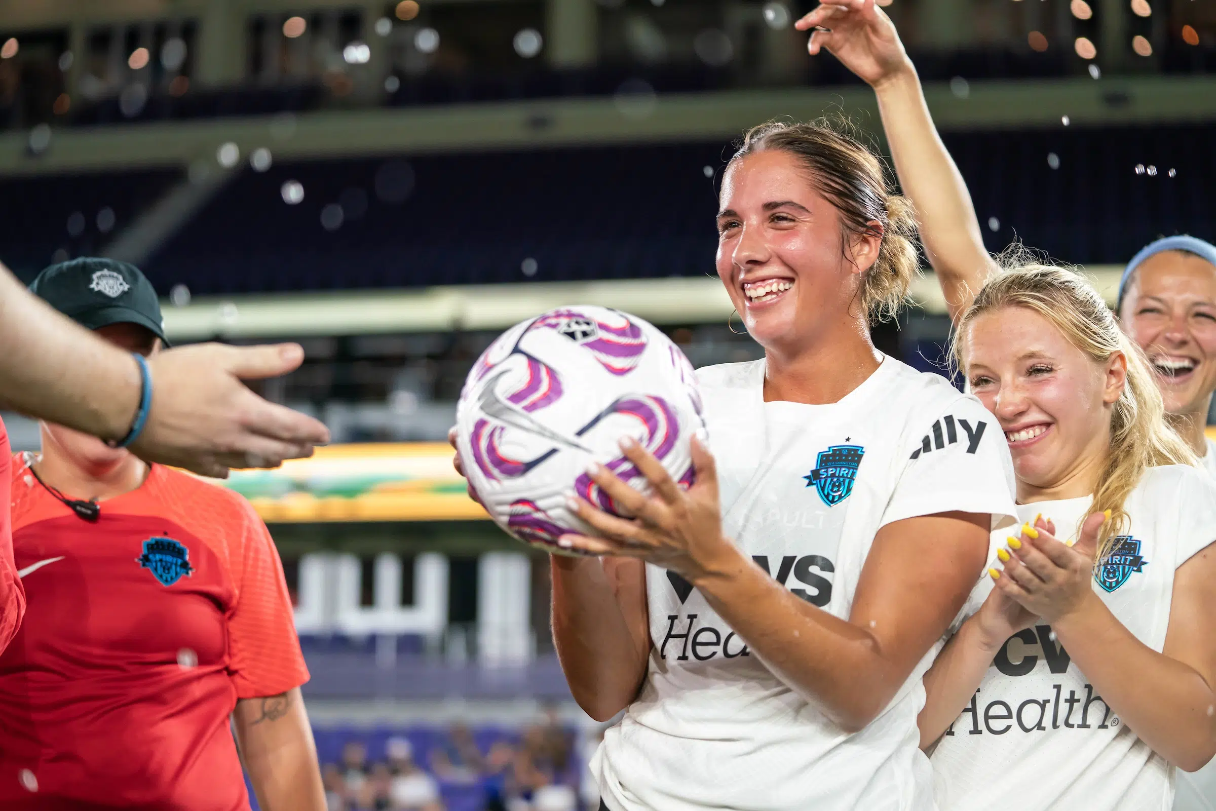 Mariana Speckmaier in a white uniform smiles as she grabs a soccer ball from outstretched hands coming in from the left side of the frame.