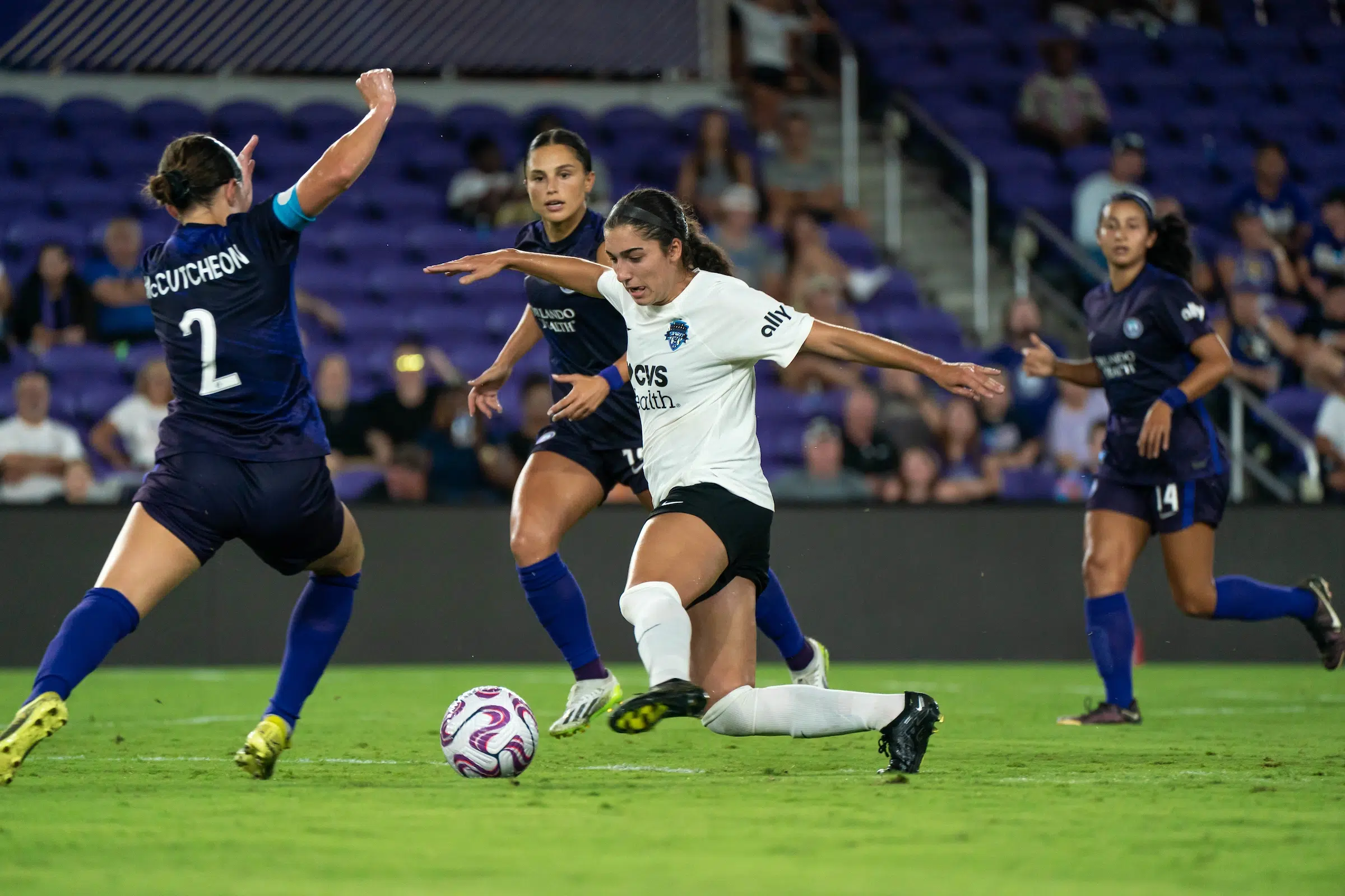 Lena Silano in a white top and black shorts attempts to dribble a soccer ball through two defenders wearing dark purple uniforms.