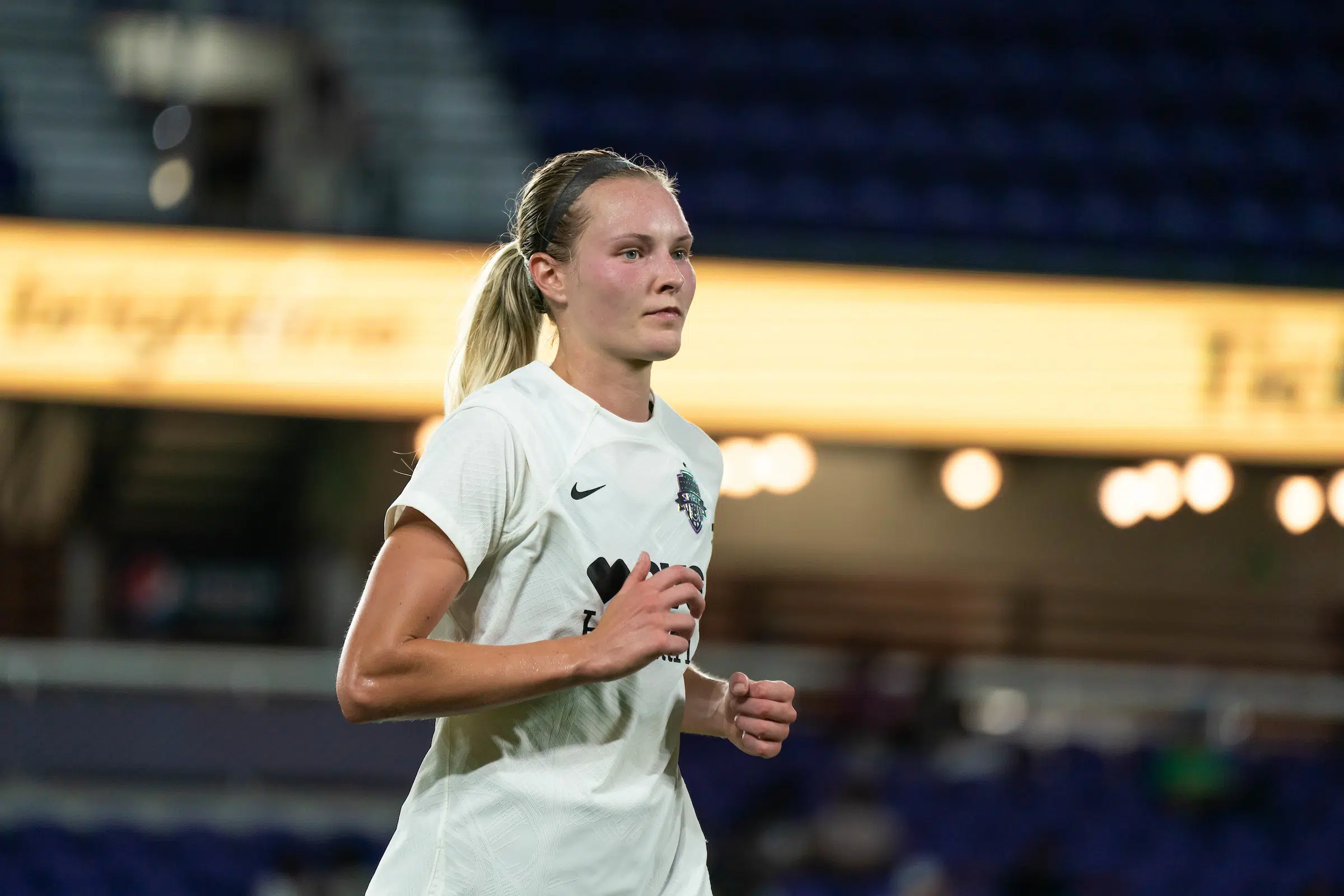 A soccer player in a white uniform with a blonde ponytail runs on a soccer field.