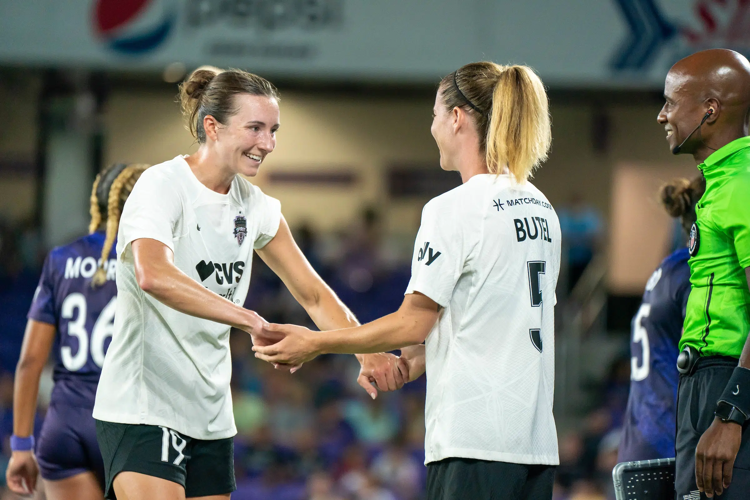 Two soccer players in white tops and black shorts shake hands as they walk off a field.
