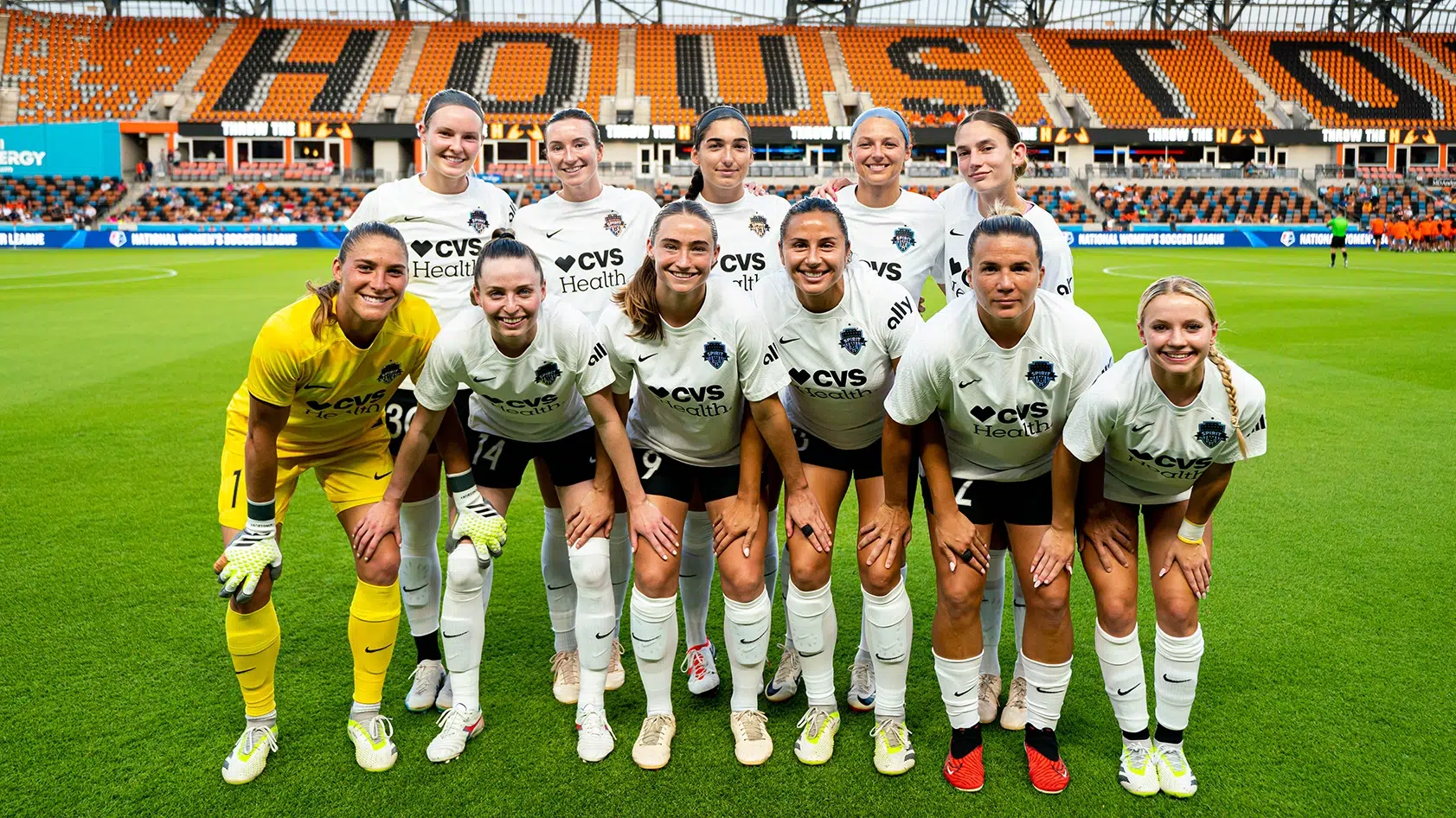 10 soccer players in white tops, black shorts and one goalie in all yellow pose for a starting eleven photo in two rows. In the background, the stadium's orange seats have 