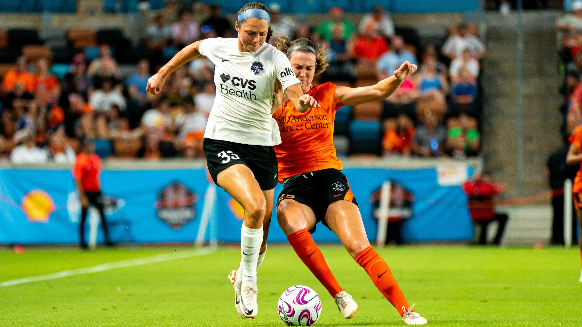 Ashley Hatch in a white top, black shorts, and white socks battles against a defender in an orange top, black shorts and orange socks for possession of a soccer ball.