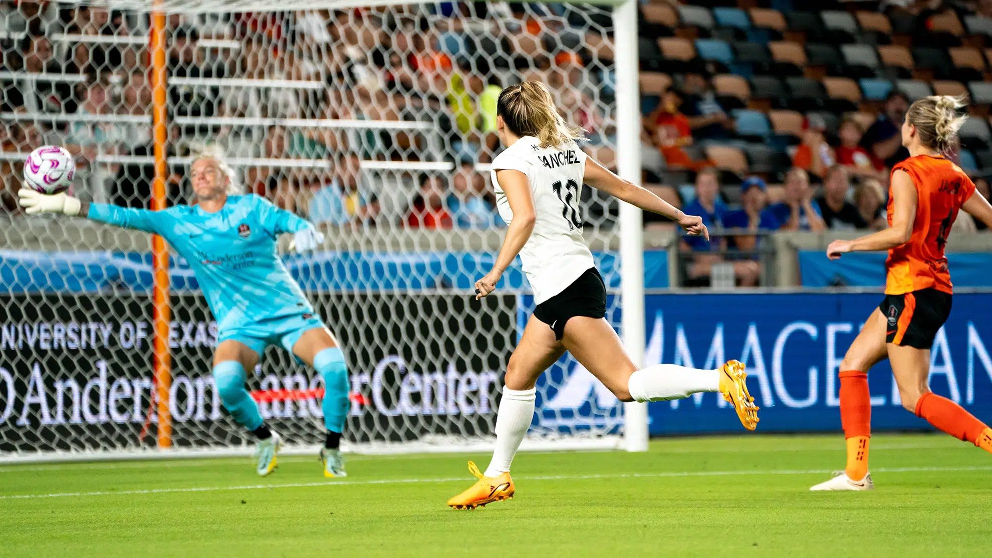 Ashley Sanchez in a white top, black shorts, white socks and orange cleats watches the soccer ball she kicked fly towards the goal. Jane Campbell in a bright blue goal uniform stretches out her right arm to save it but is just a bit short.