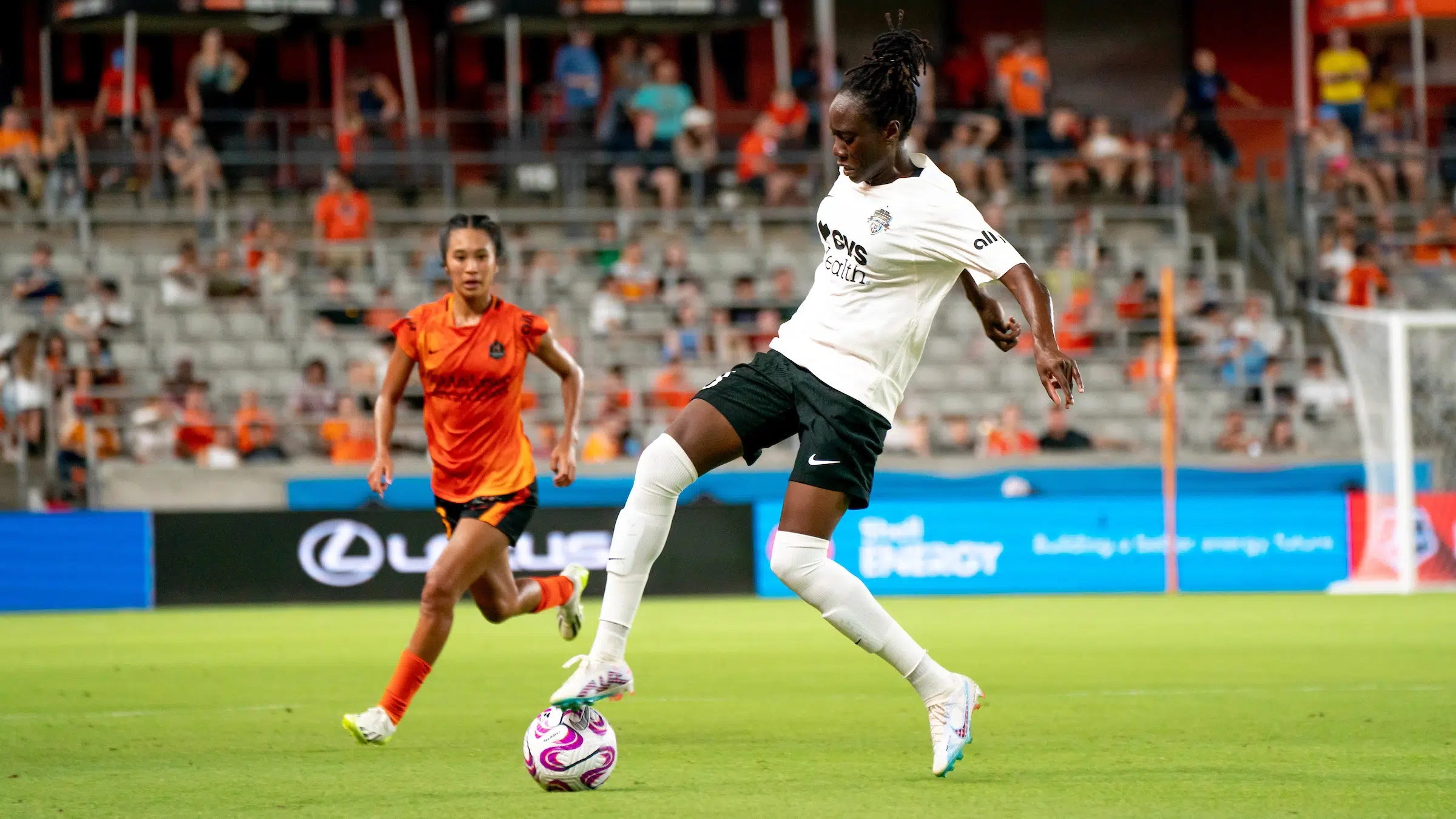 Ouleymata Sarr in a white shirt, black shorts, white socks and white cleats controls a soccer ball with her right foot as a defender in an orange uniform tries to challenge her.