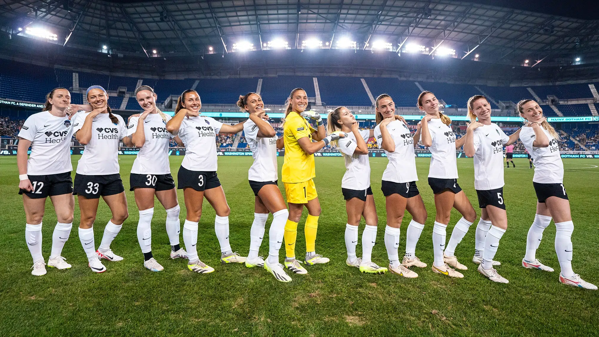 The starting eleven of the Washington Spirit pose with their hands under their chins. The team is wearing white tops, black shorts and white socks. Aubrey Kingsbury is in the middle wearing a yellow goalkeeper jersey.