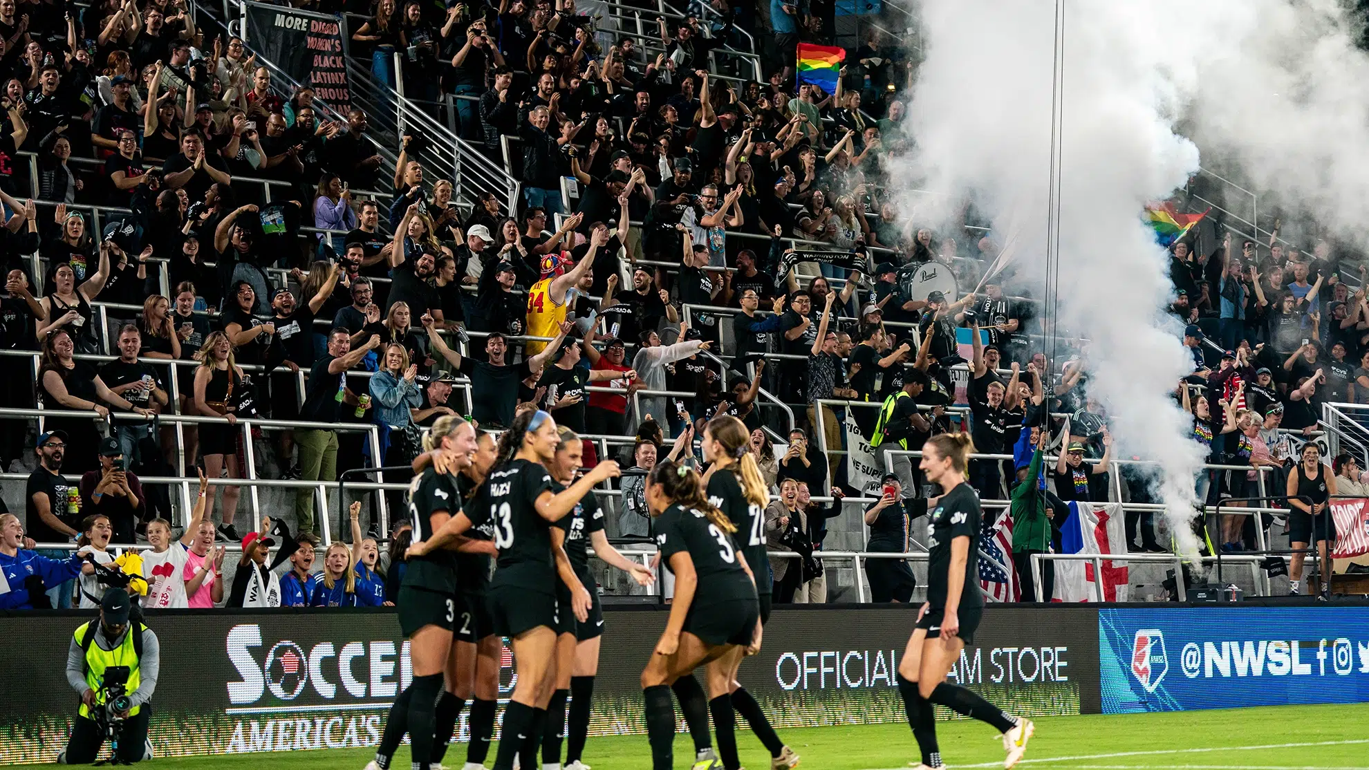 In the foreground, Spirit players wearing black celebrate. In the background, supporters in the stands raise their hands, yell and release smoke in celebration.