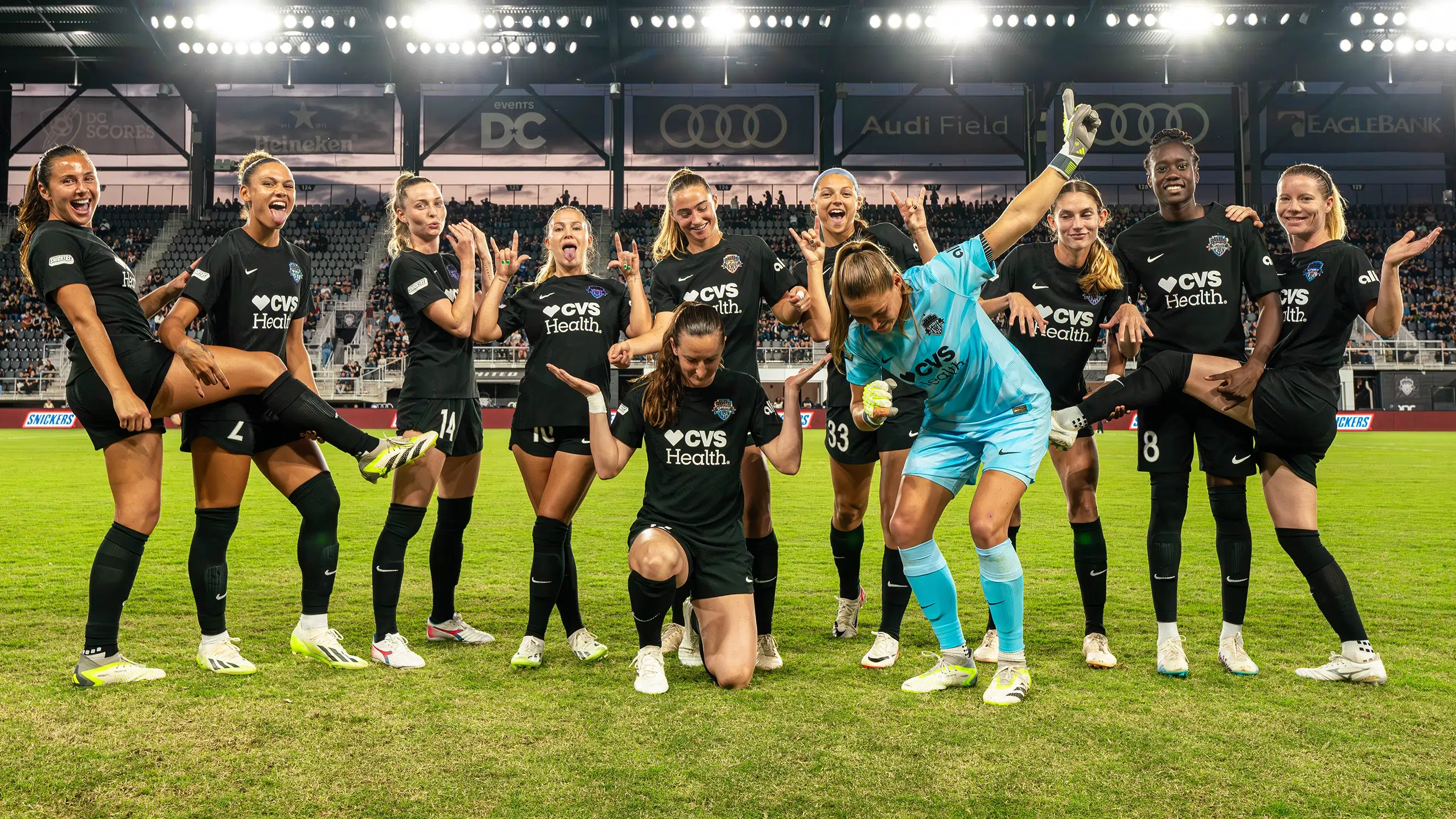 The starting XI for the Washington Spirit pose in different rock and roll stances. They wear all black uniforms except the goalie in light blue.