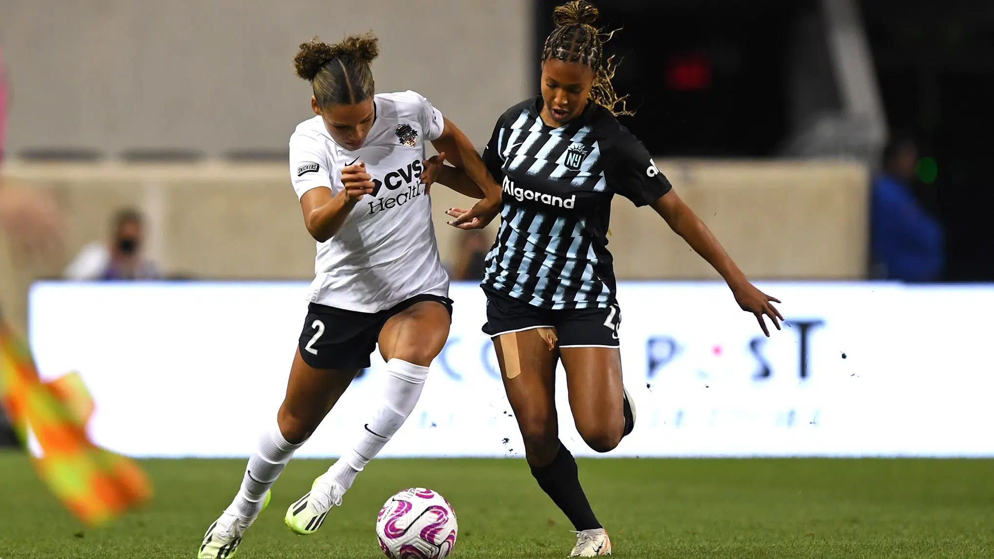 Trinity Rodman in a white jersey with black shorts battles Midge Purce in a black jersey and shorts for possession of the soccer ball.