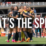 The Washington Spirit huddle and hug after scoring a goal at Audi Field. White text reading "That's the Spirit" is overlayed on the image.