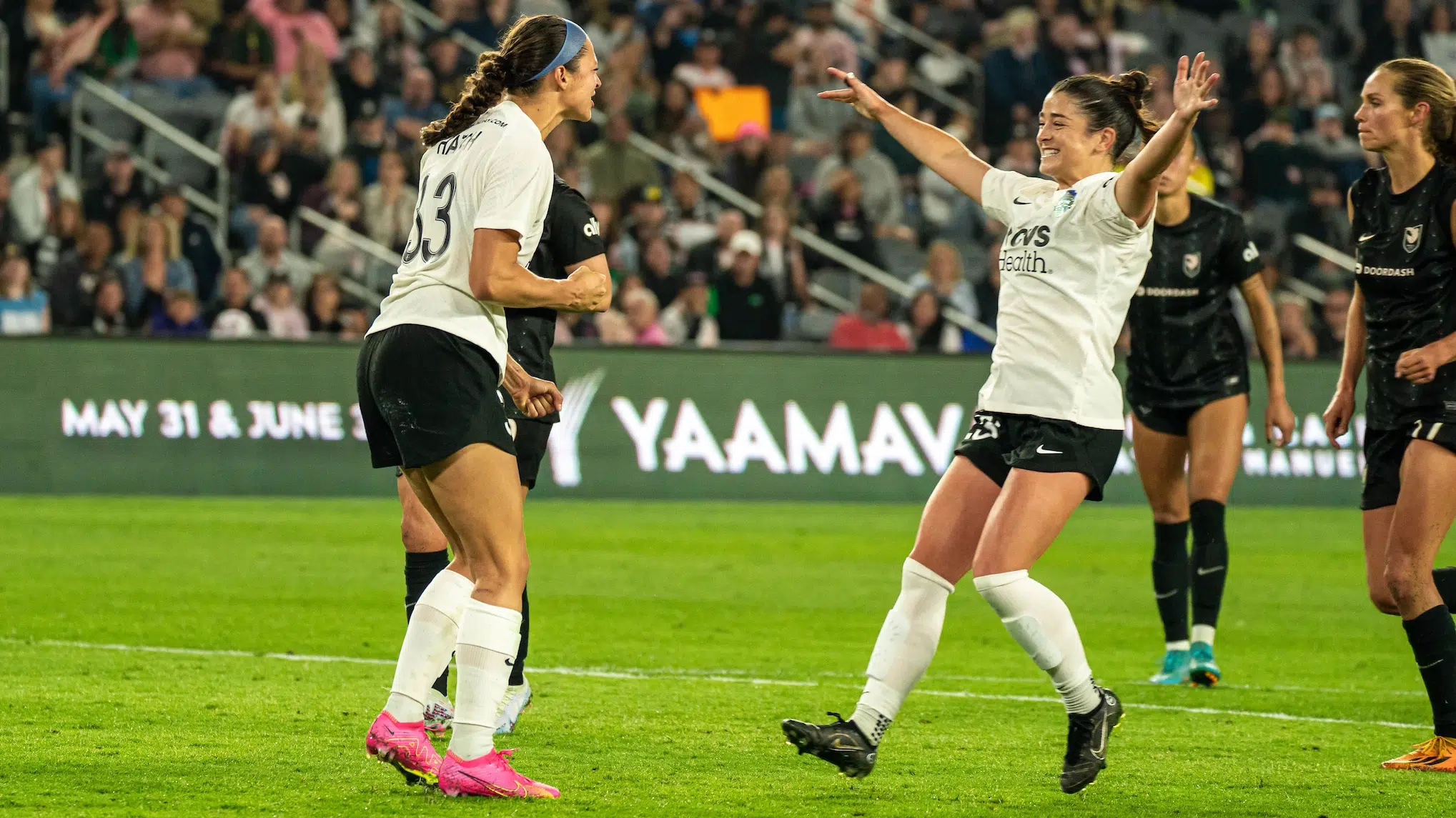 Ashley Hatch in a white top, black shorts and pink cleats pumps her fist in celebration as Marissa Sheva in a white top, black shorts and black cleats runs towards her with her arms wide open smiling.