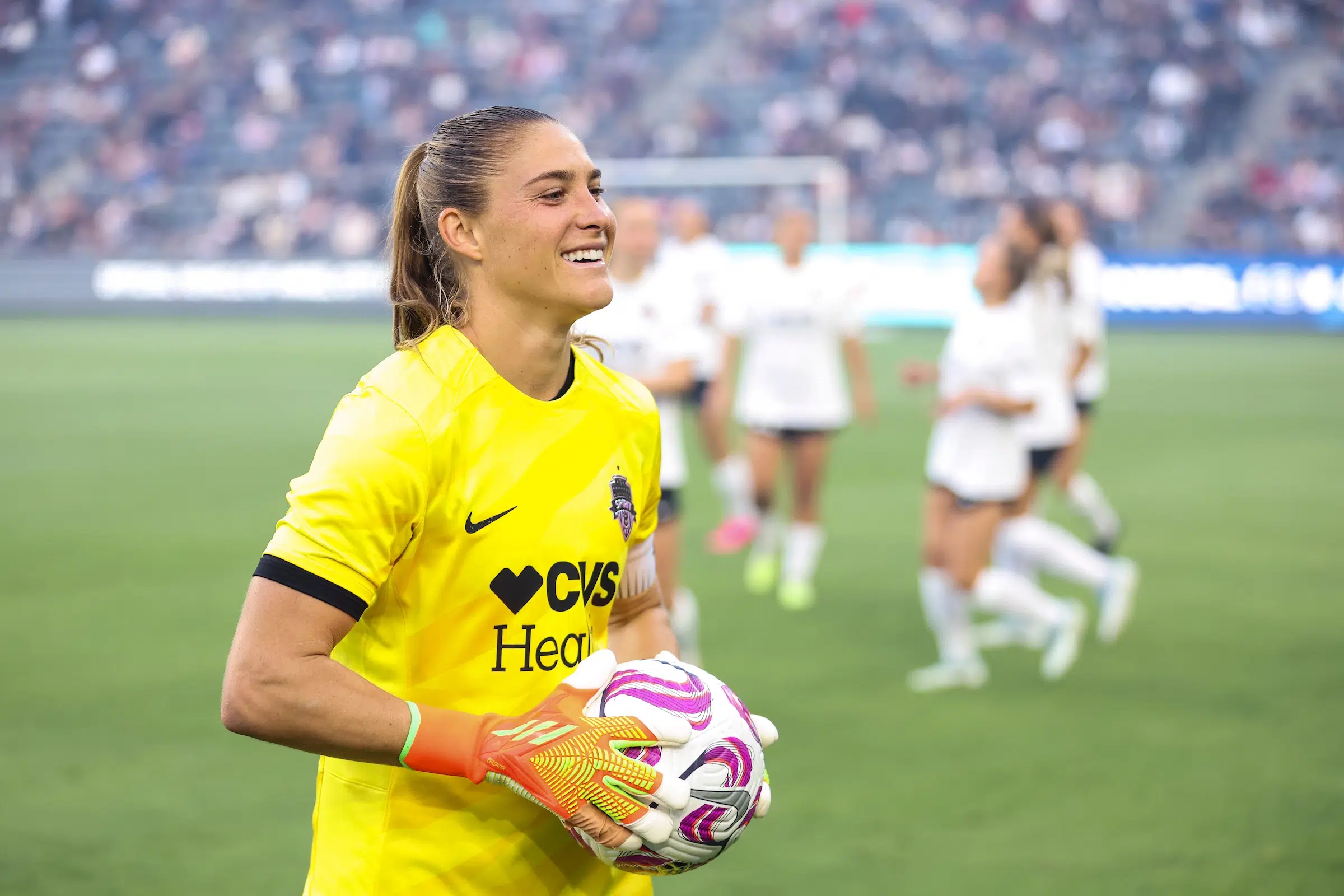 Ashley Hatch in a yellow uniform smiles as she holds a soccer ball in two hands.