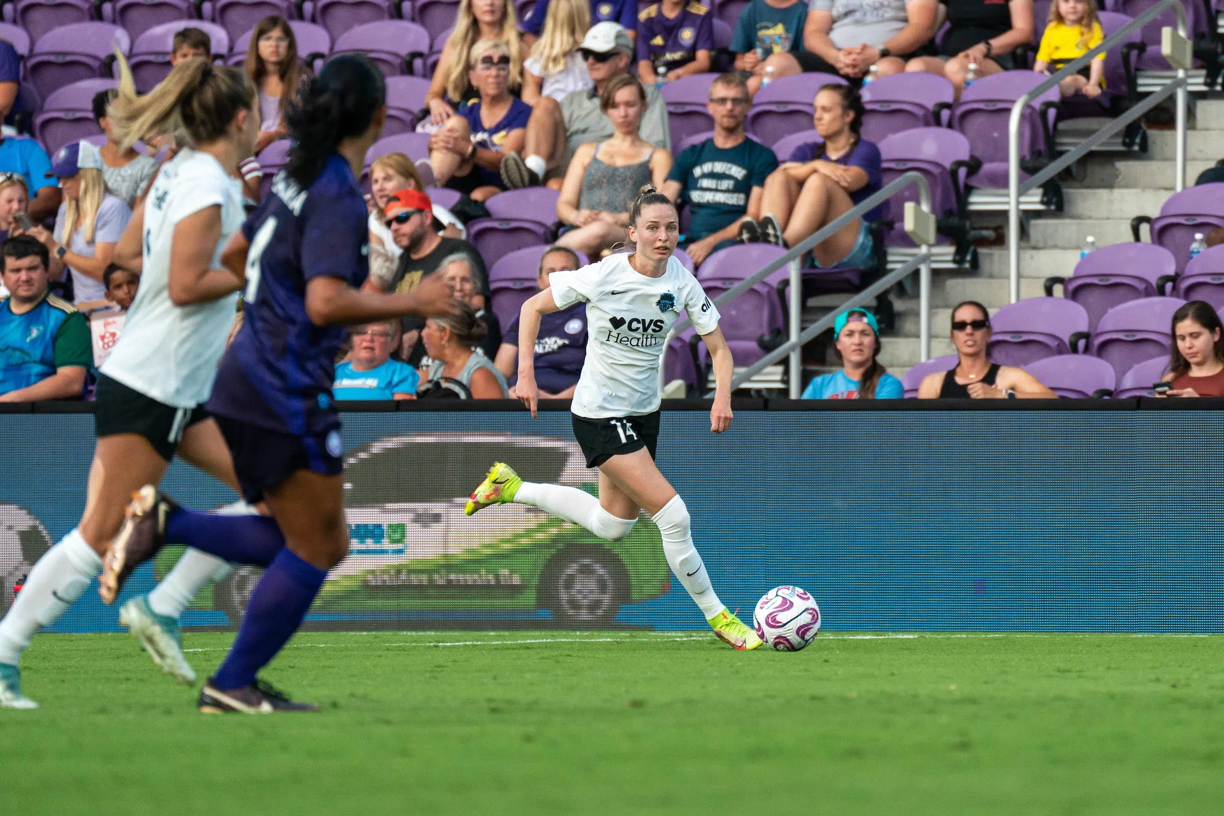 Gabby Carle in a white top, black shorts and white socks dribbles a soccer. In the foreground, two players are running out of focus.