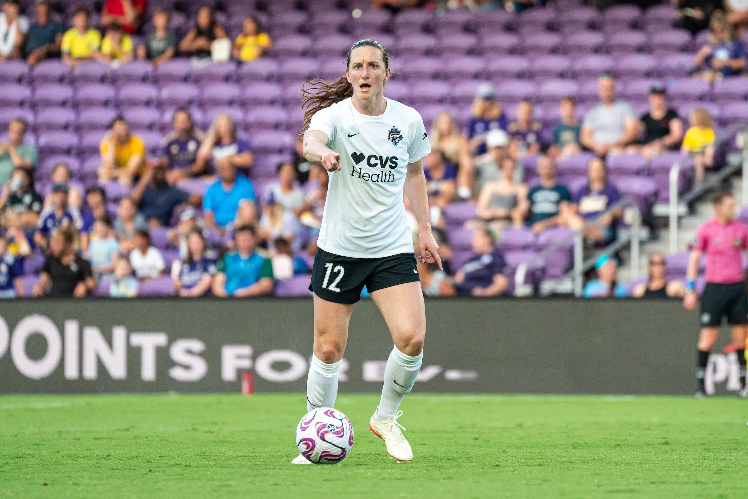 Andi Sullivan in a white top, black shorts and white socks dribbles a soccer ball and points with the index finger of her right hand.