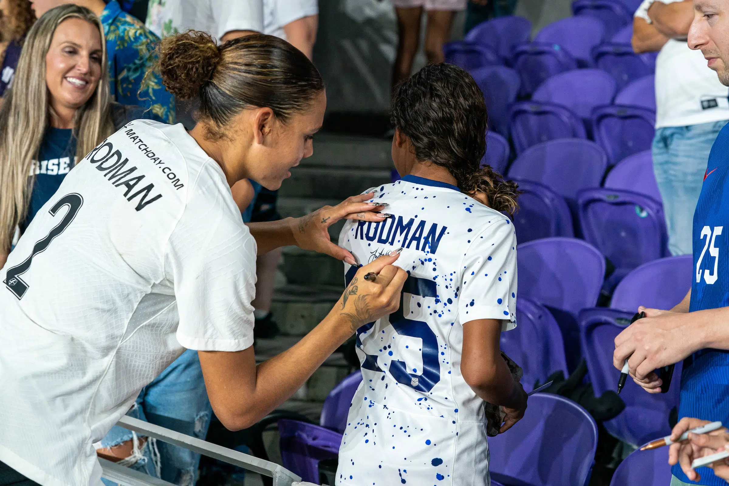 Trinity Rodman in a white top signs the back of a USWNT Rodman #25 jersey worn by a young fan.