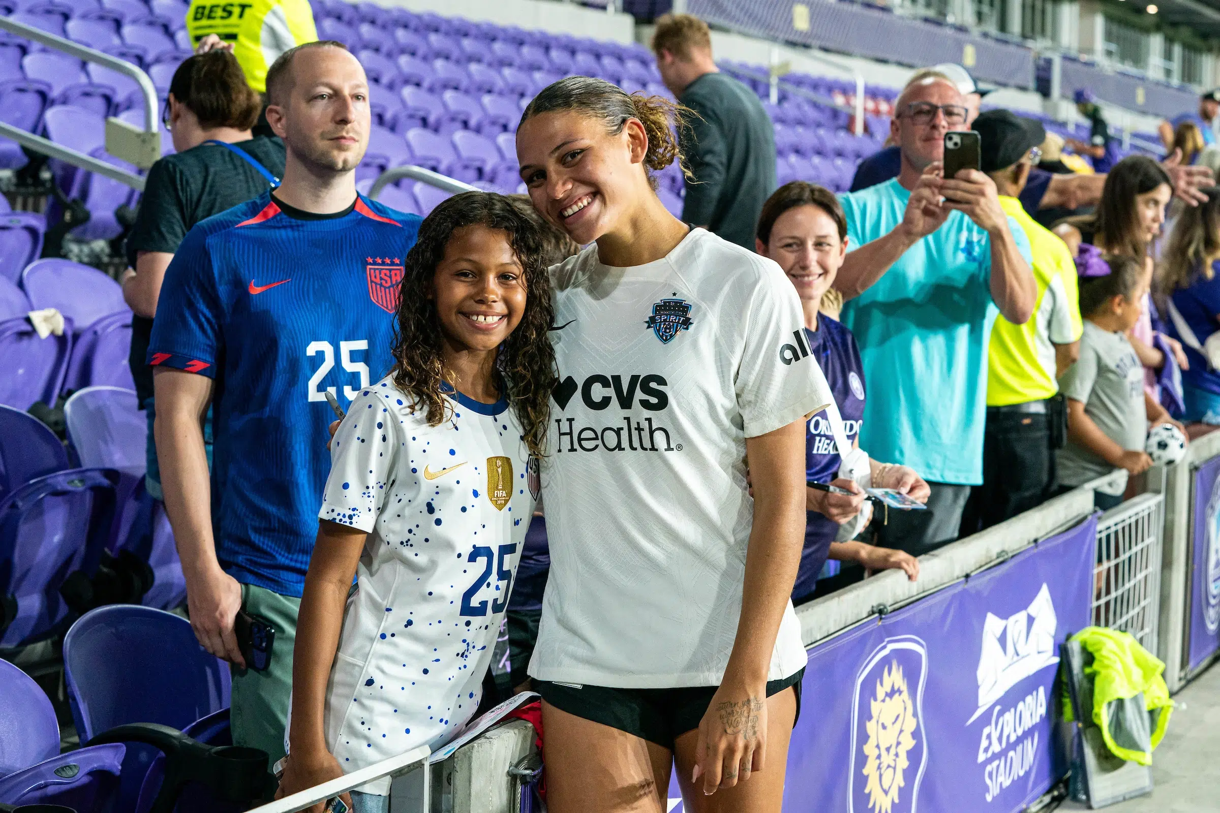 Trinity Rodman smiles and poses with a young fan. Rodman is wearing a white top and black shorts. The young fan is wearing a white USWNT jersey speckled with blue.