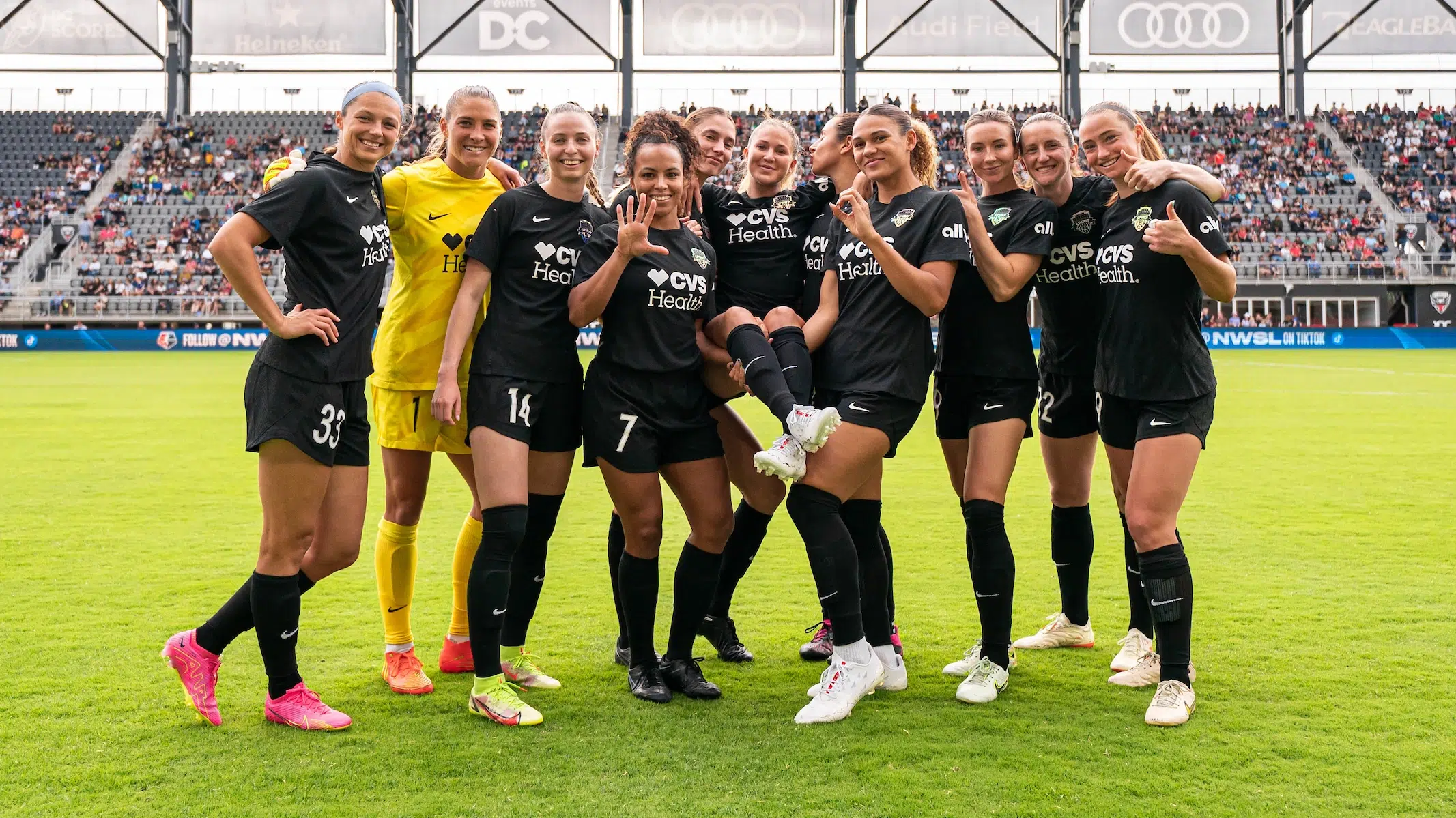 Ashley Sanchez is held up by two of her teammates as seven others circle around and hold up the number 50 in celebration of her 50th cap. Ten of the players are in black uniforms and one is in a yellow goalie uniform.