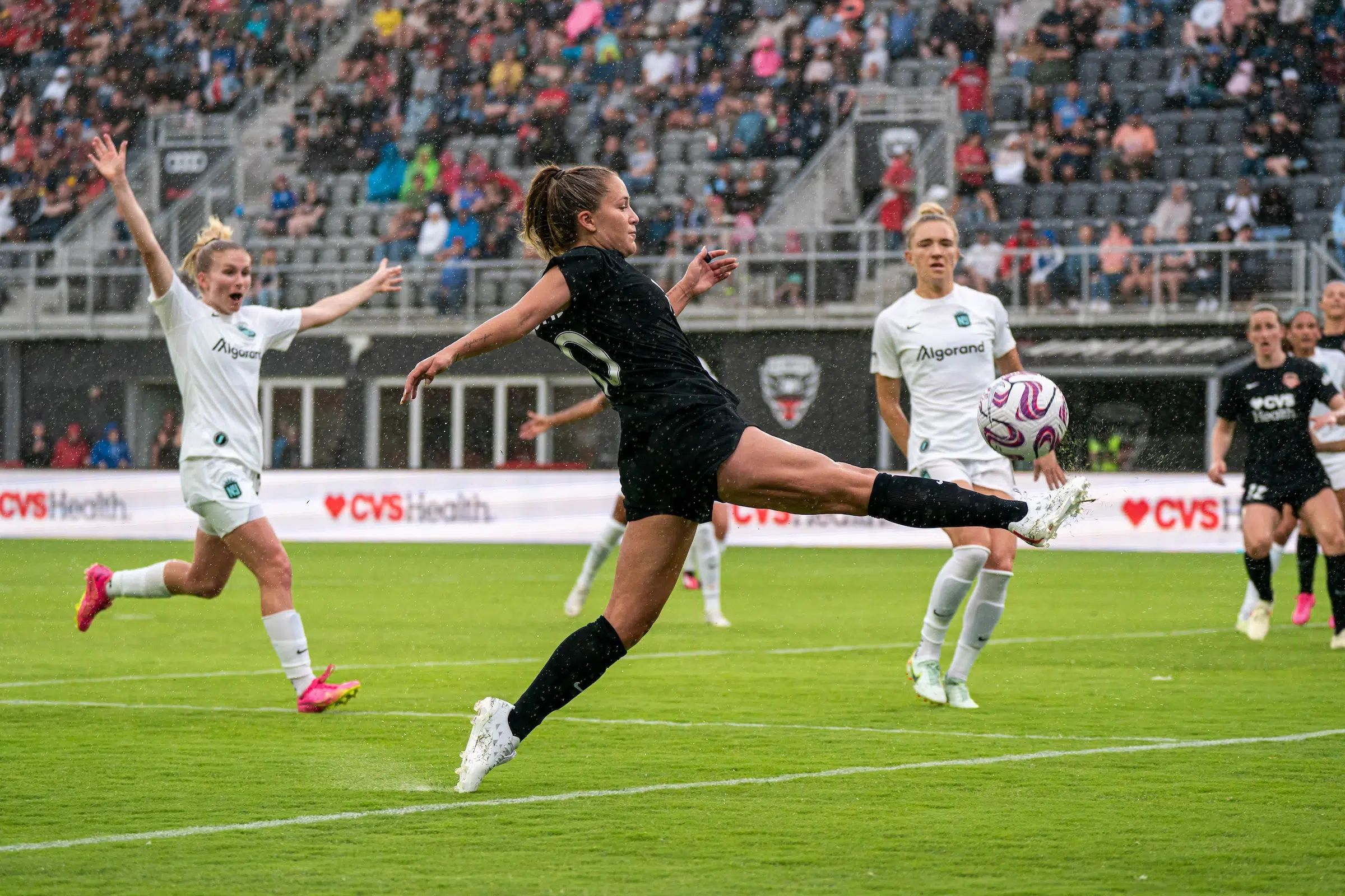 Ashley Sanchez in an all-black uniform extends her leg out to trap a soccer ball as two defenders in white jerseys look on. Rain is visibly pouring down onto the field.