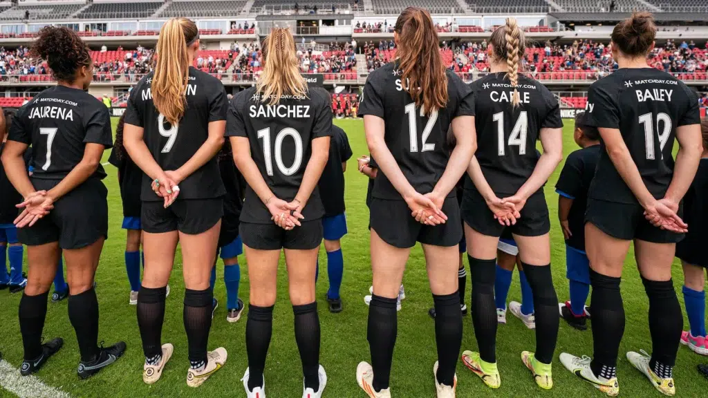 Six soccer players in black uniforms stand facing away from the camera with their hands clasped behind their backs.