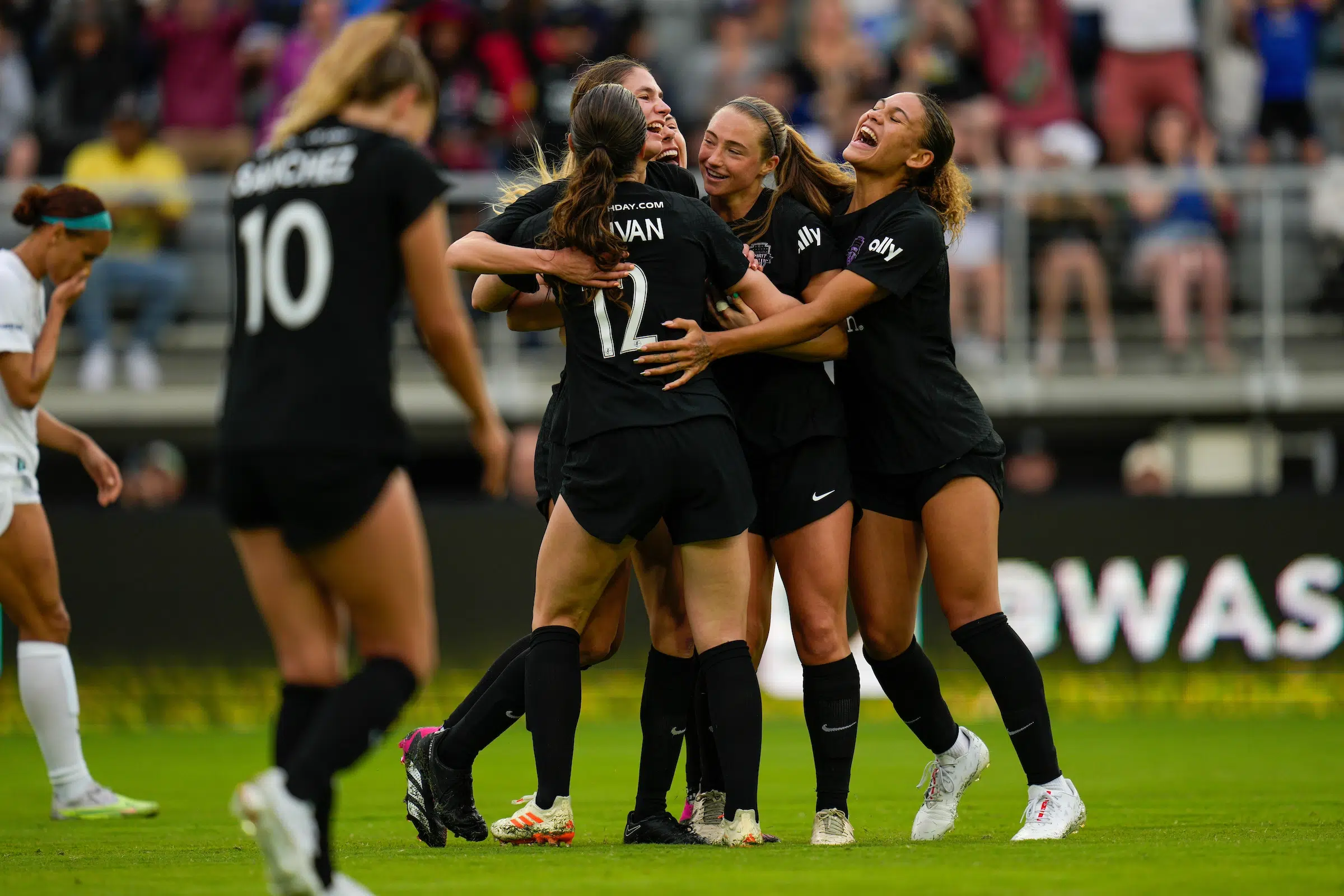 Six soccer players in black uniforms smile and hug in celebration after a goal.