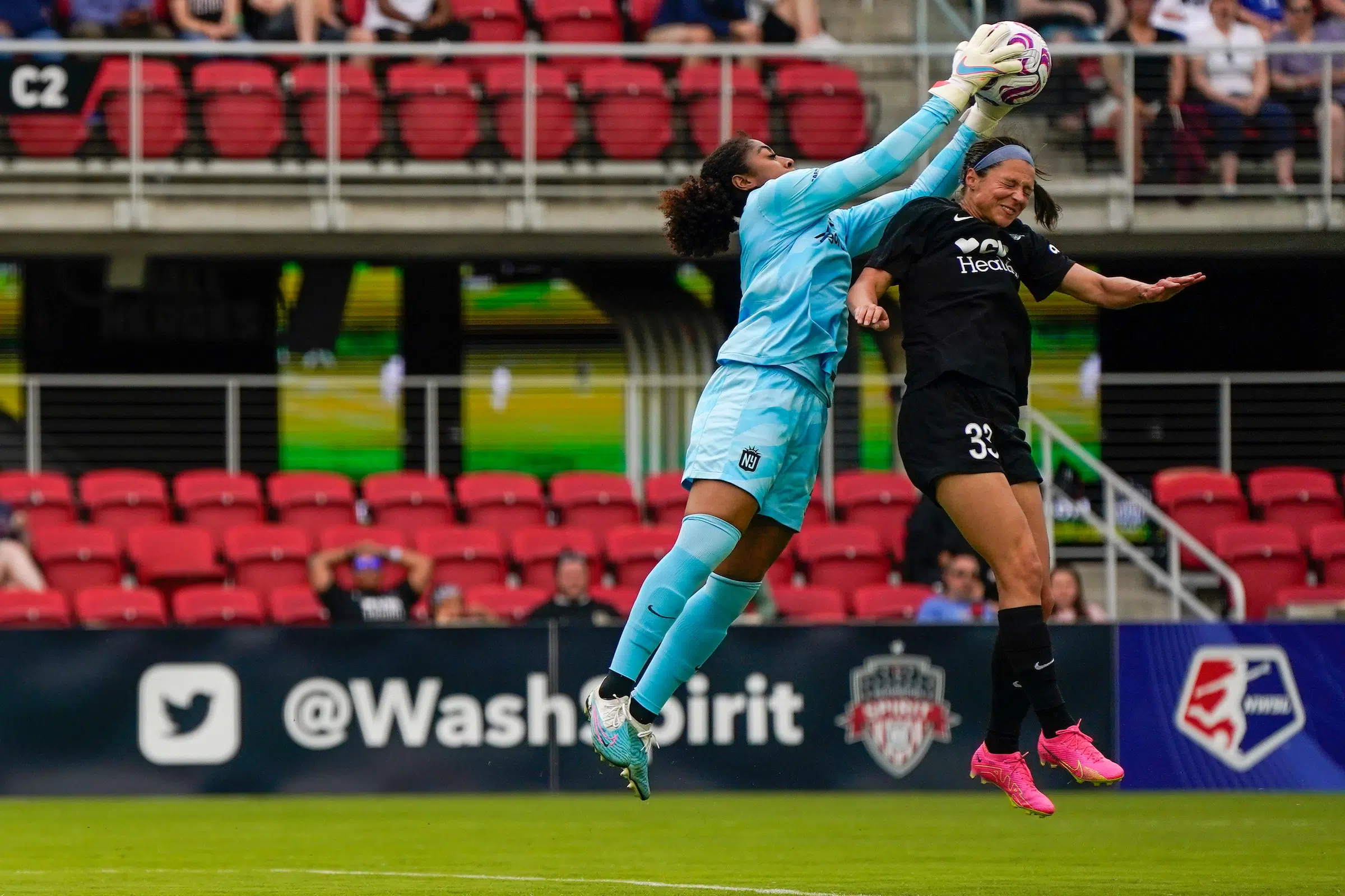 Ashley Hatch in a black uniform and pink cleats jumps to try and head a soccer ball as a goalie in a bright blue uniform jumps up to grab the ball.
