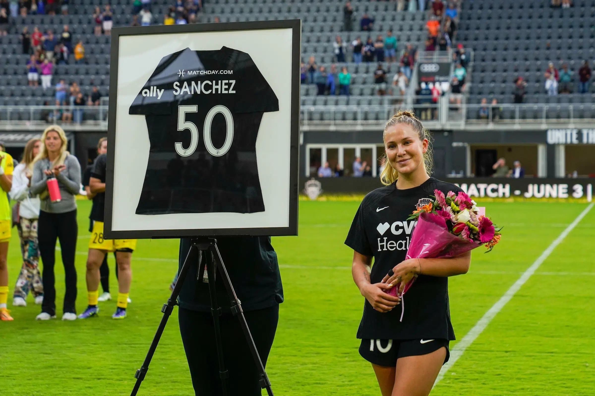 Ashley Sanchez in a black uniform holds a bouquet in her arms as she poses next to a framed black jersey with the name Sanchez and the number 50 to celebrate her 50 NWSL caps.