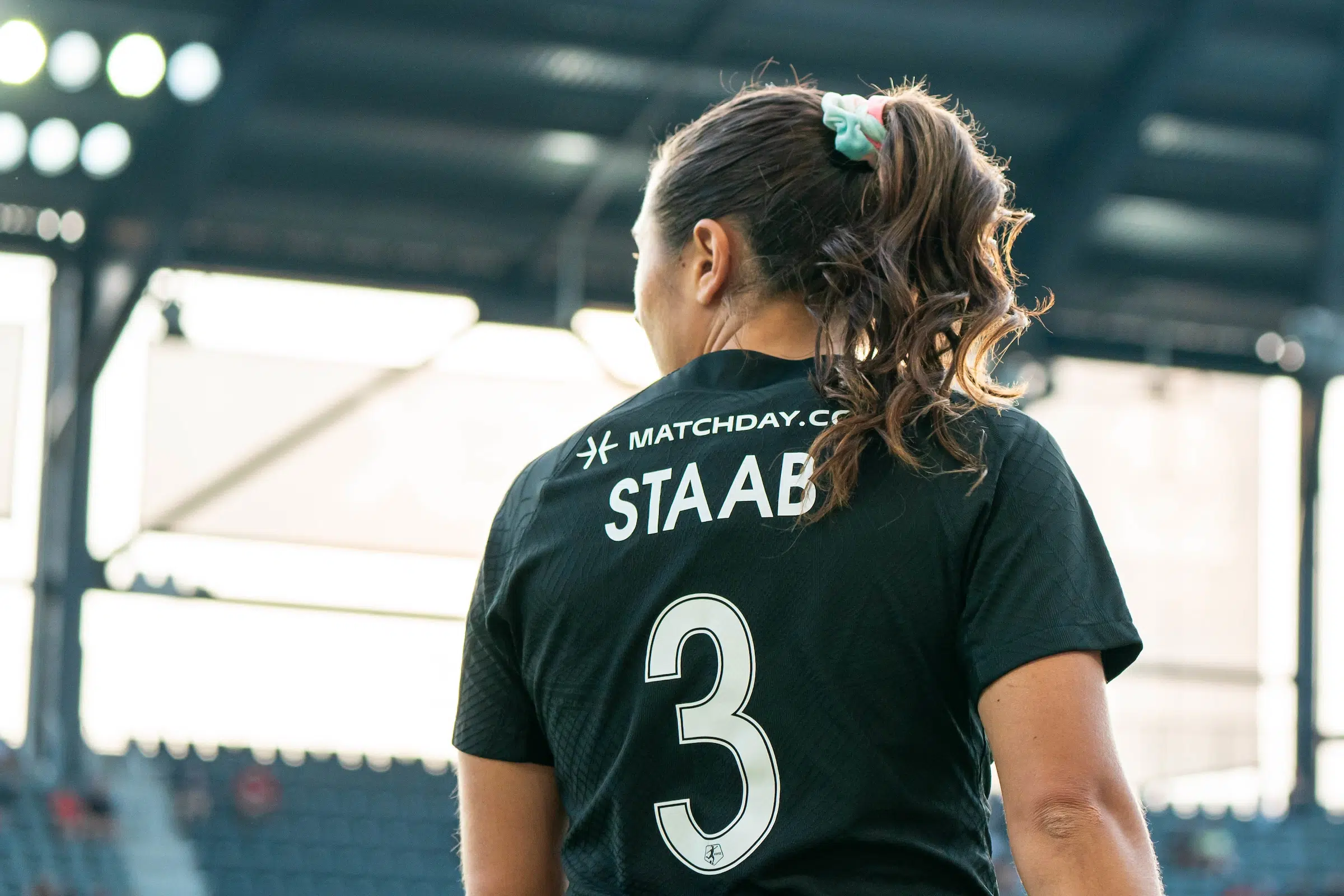 Sam Staab in a black jersey looks off to her left. Her curly ponytail is held up by a multi-colored scrunchie.