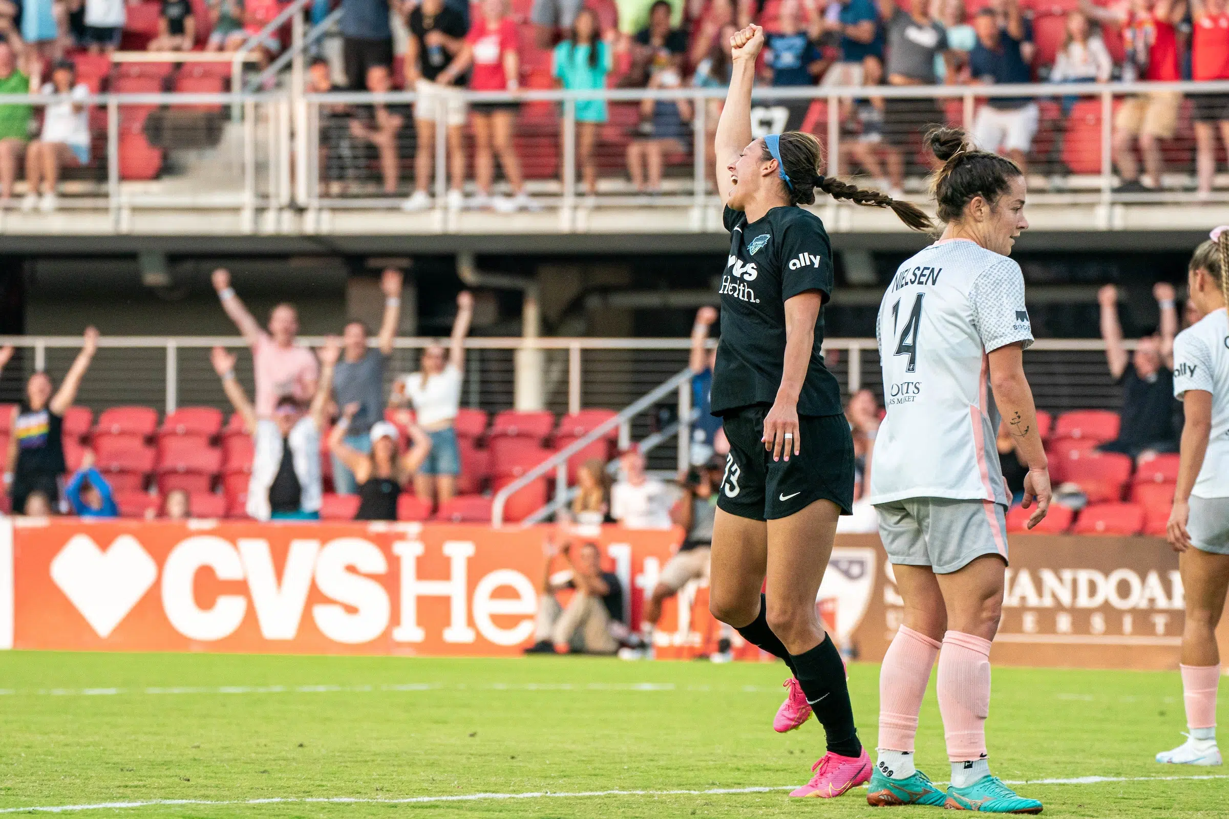 Ashley Hatch in a black uniform with pink cleats lifts her fist in celebratio as two defenders in gray uniforms look on in disappointment.