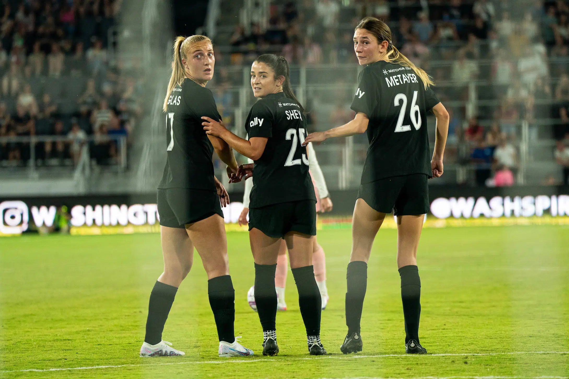 Three players in black uniforms stand in a line shoulder-to-shoulder and look back towards their goalie.