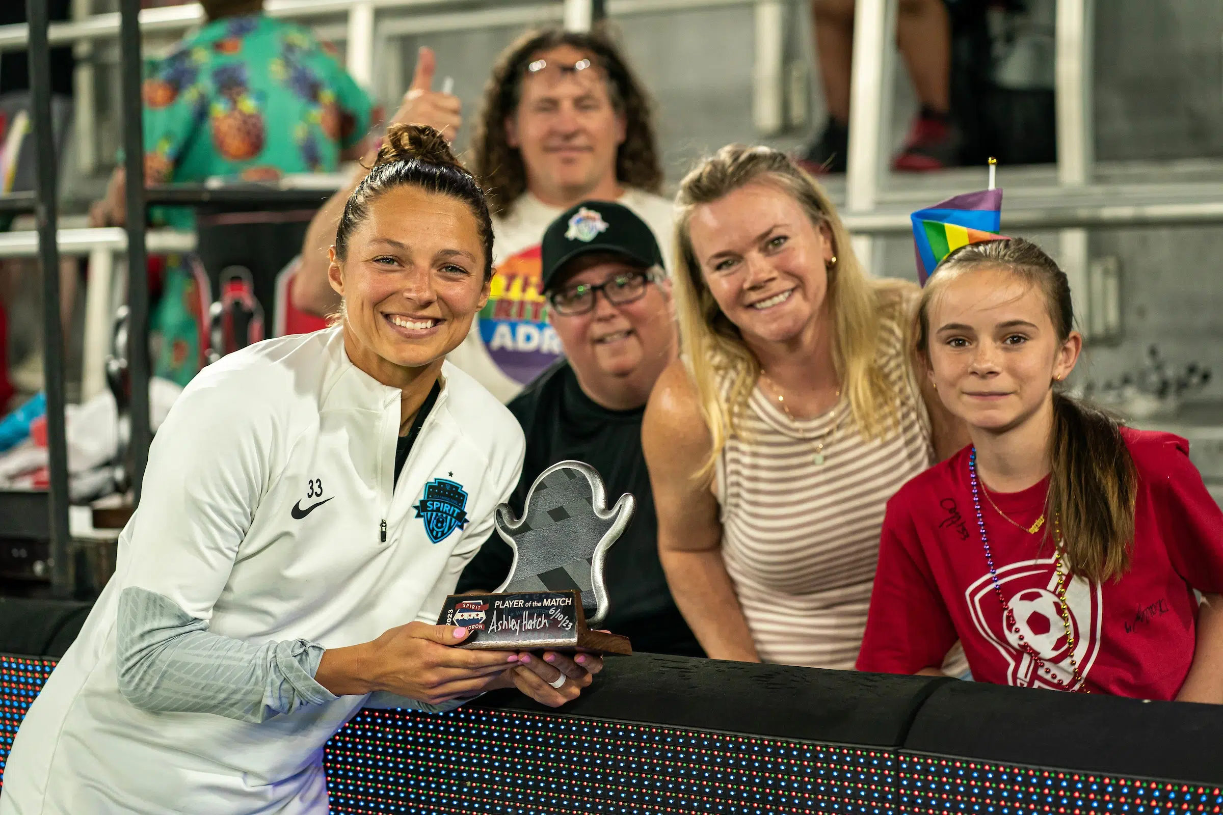 Ashley Hatch in a white quarter zip top holds a ghost-emoji shaped trophy and poses for a photo with a young fan.