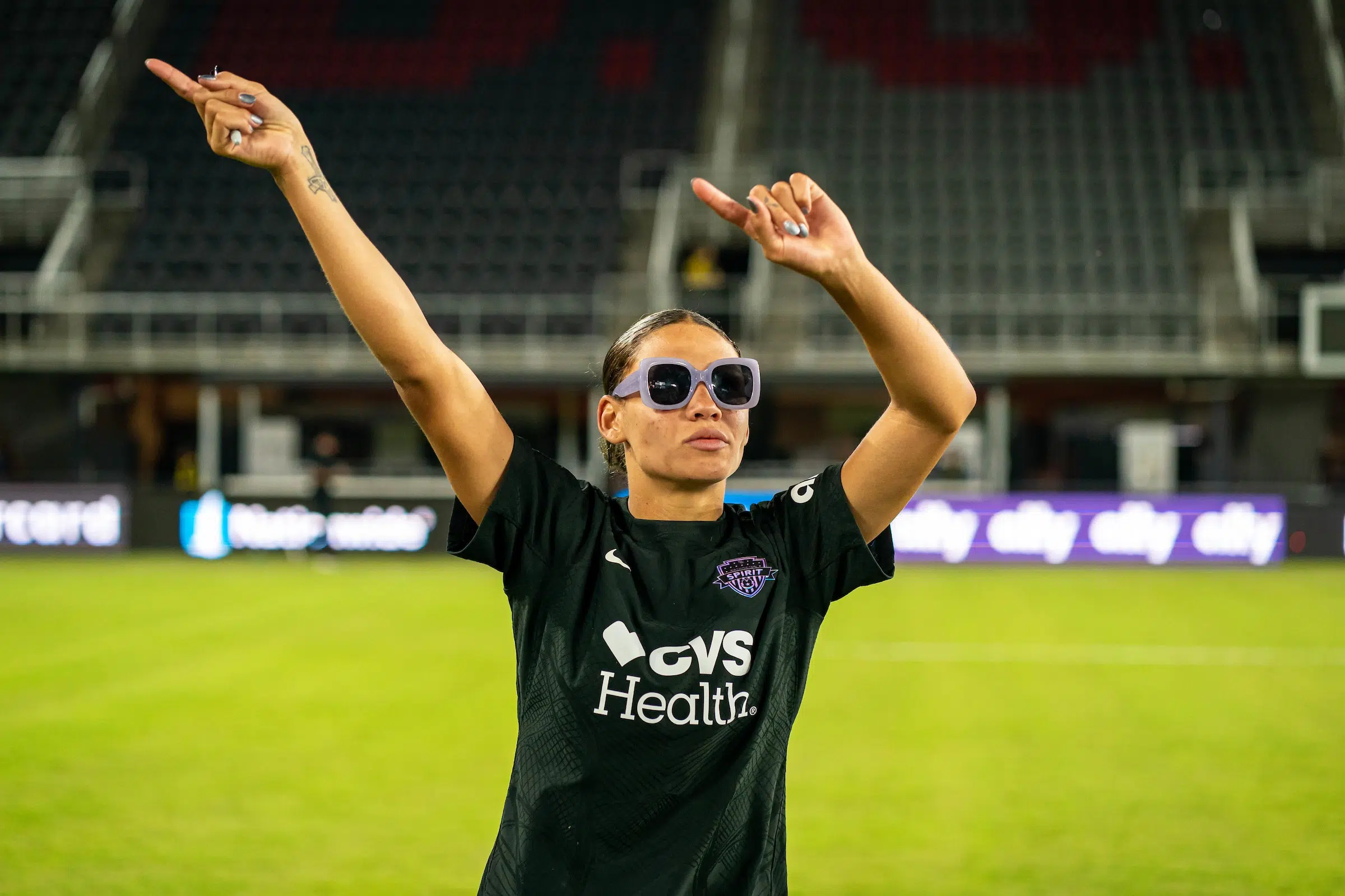 Trinity Rodman wears a black jersey and purple sunglasses as she dances with both hands in the air.