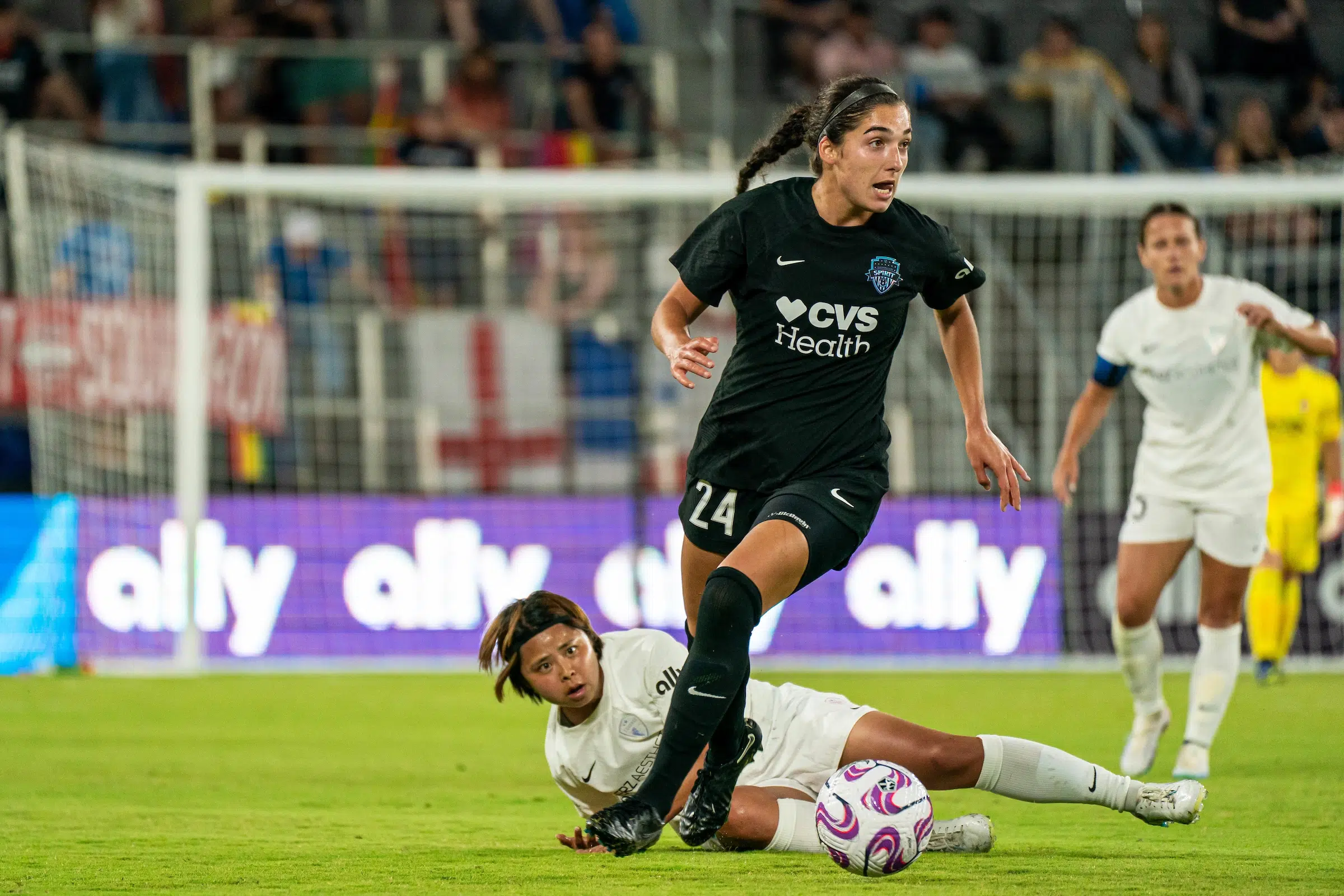 Lena Silano in a black uniform dribbles the soccer ball past a defender in a white uniform who is lying on the ground.