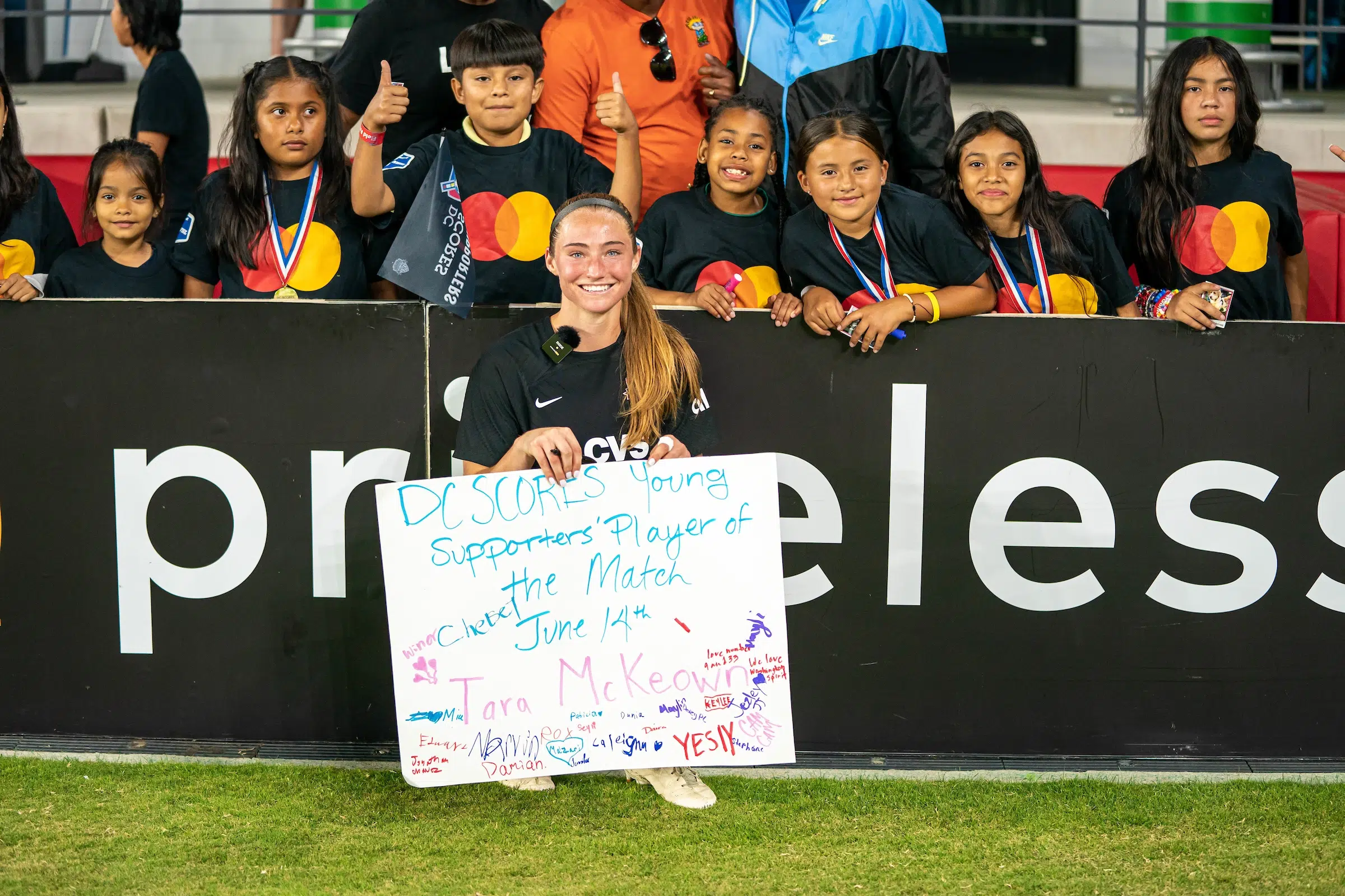 Tara McKeown poses for a photo holding a poster made by the young fans in the photo who all wear black tshirts.