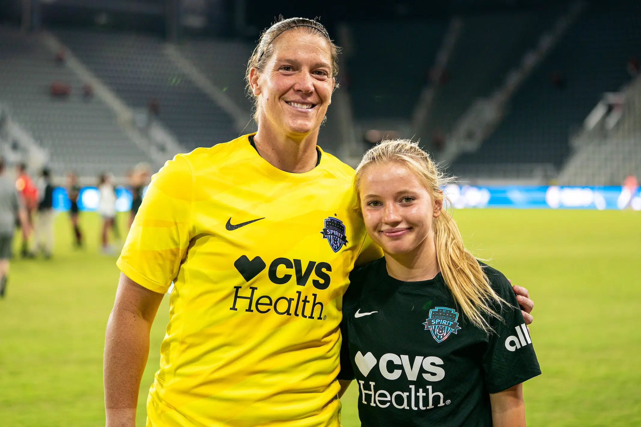 Nicole Barnhart in a yellow goalie jersey and Chloe Ricketts in a black jersey have their arms around each other and smile. Barnhart stands about a foot taller.