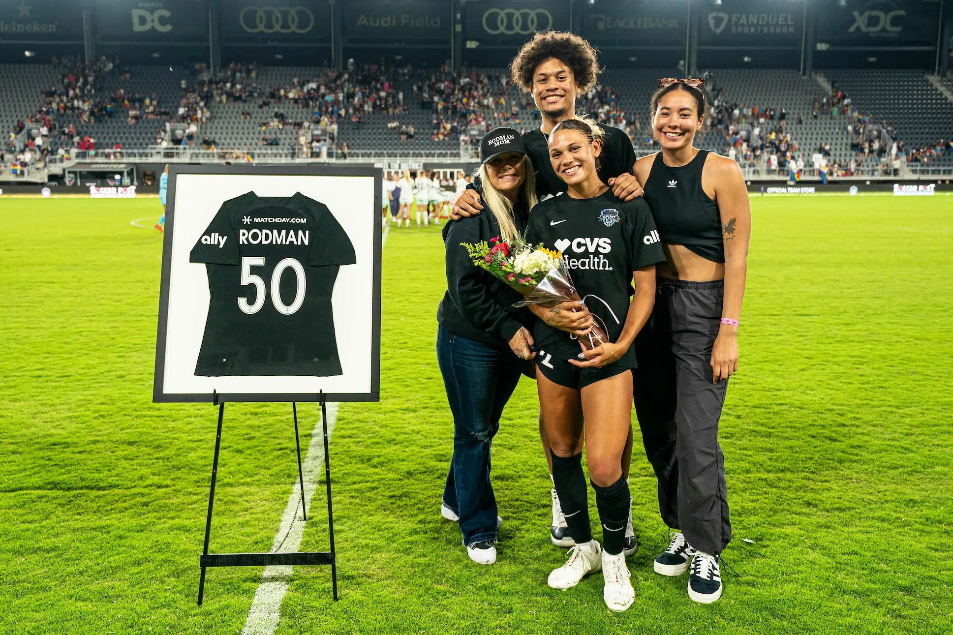 Trinity Rodman in a black uniform holds a bouquet of flowers and is surrounded by her mother, brother and friend as they take a photo next to a framed black jersey with her name and the 50 on it to celebrate her 50 caps.