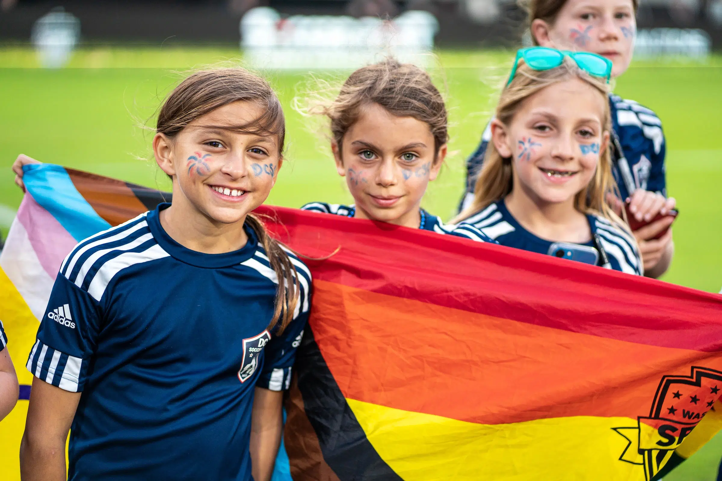 Three young fans in navy blue soccer jerseys hold up an LGBTQ+ flag.