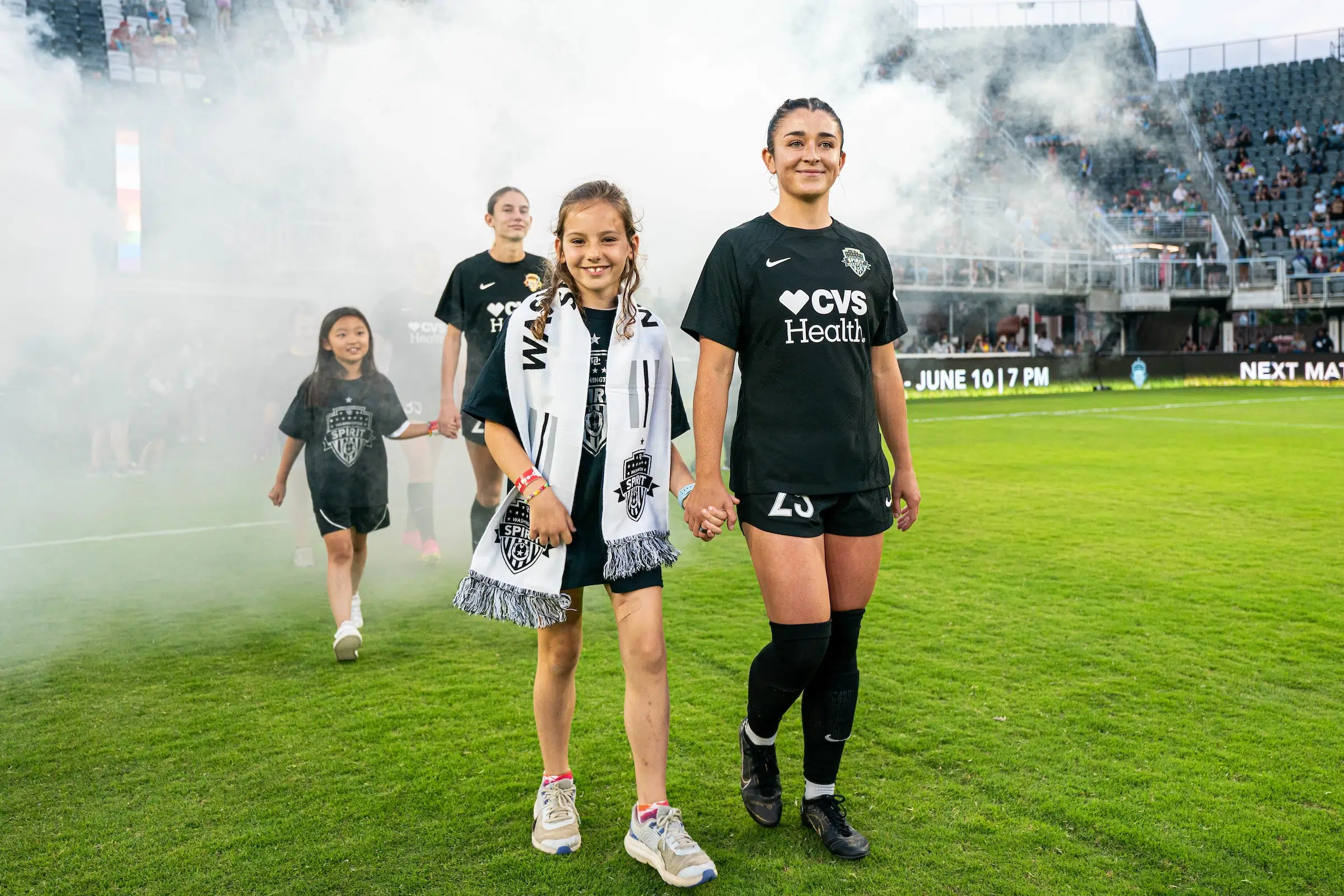Marissa Sheva in an all black uniforms walks out of the tunnel onto the soccer field holding hands with a young fan who is wearing a Washington Spirit scarf. Behind them, smoke machines fill the air.