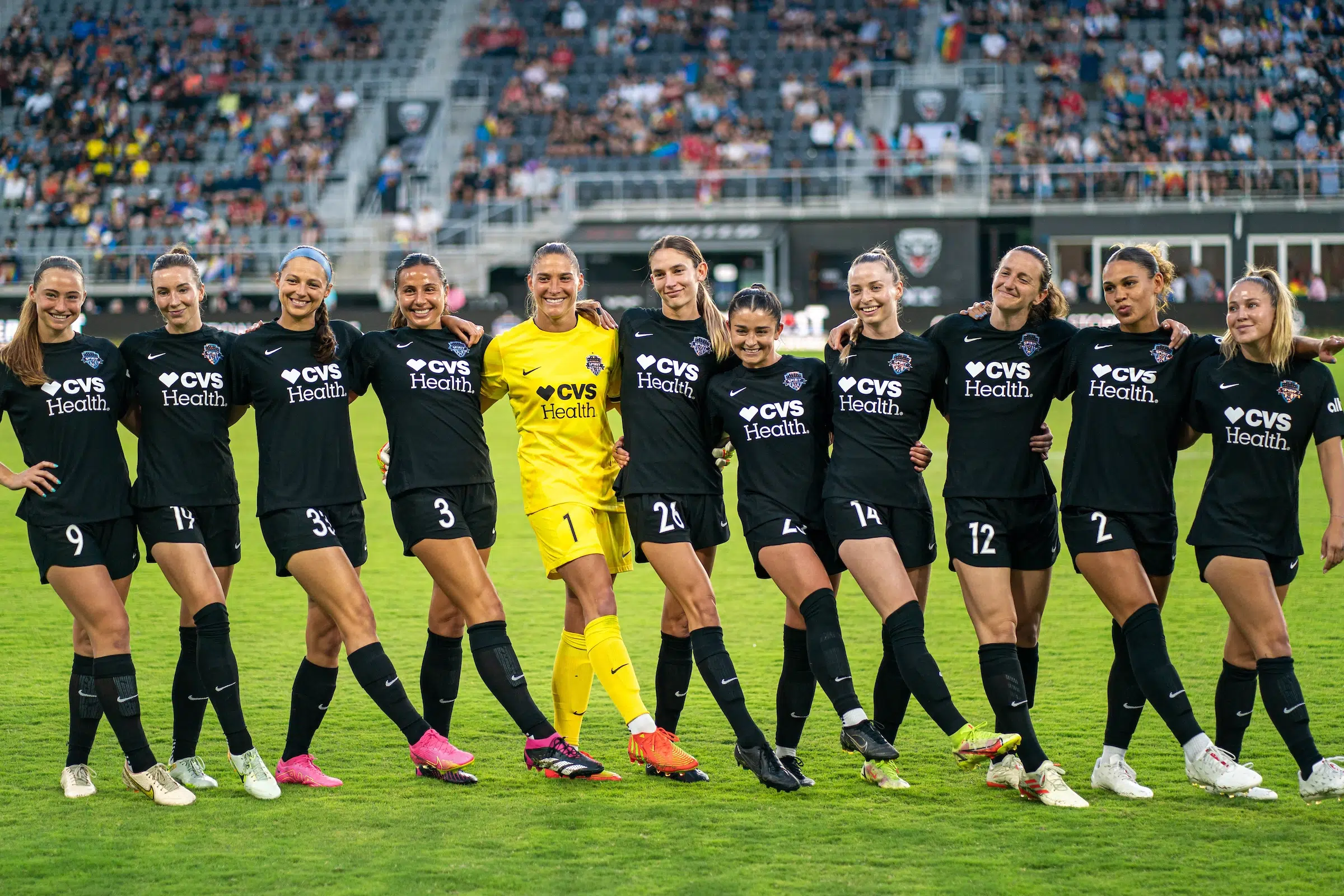 10 players in black uniforms and one in a yellow uniform line up with their arms around each other and outstretch their right legs with their toes pointed.