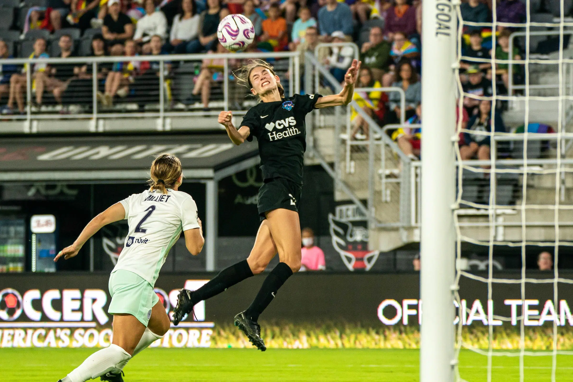Paige Metayer in a black uniform jumps up to head a soccer ball as a defender in a light colored uniform attempts to defend her.