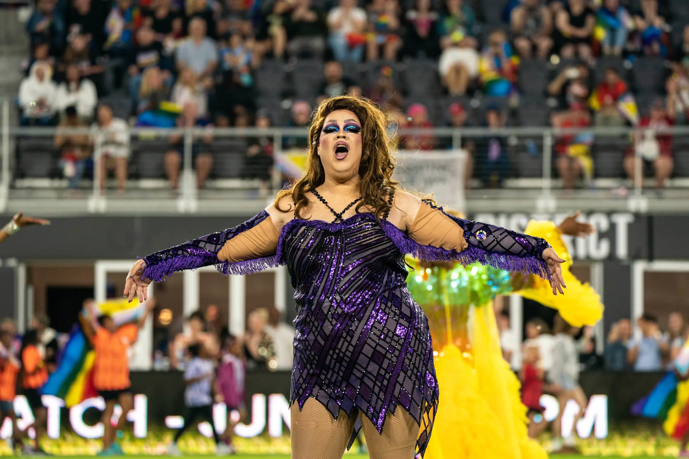 A drag queen in a purple and black sparkly dress belts out a song during a halftime performance.