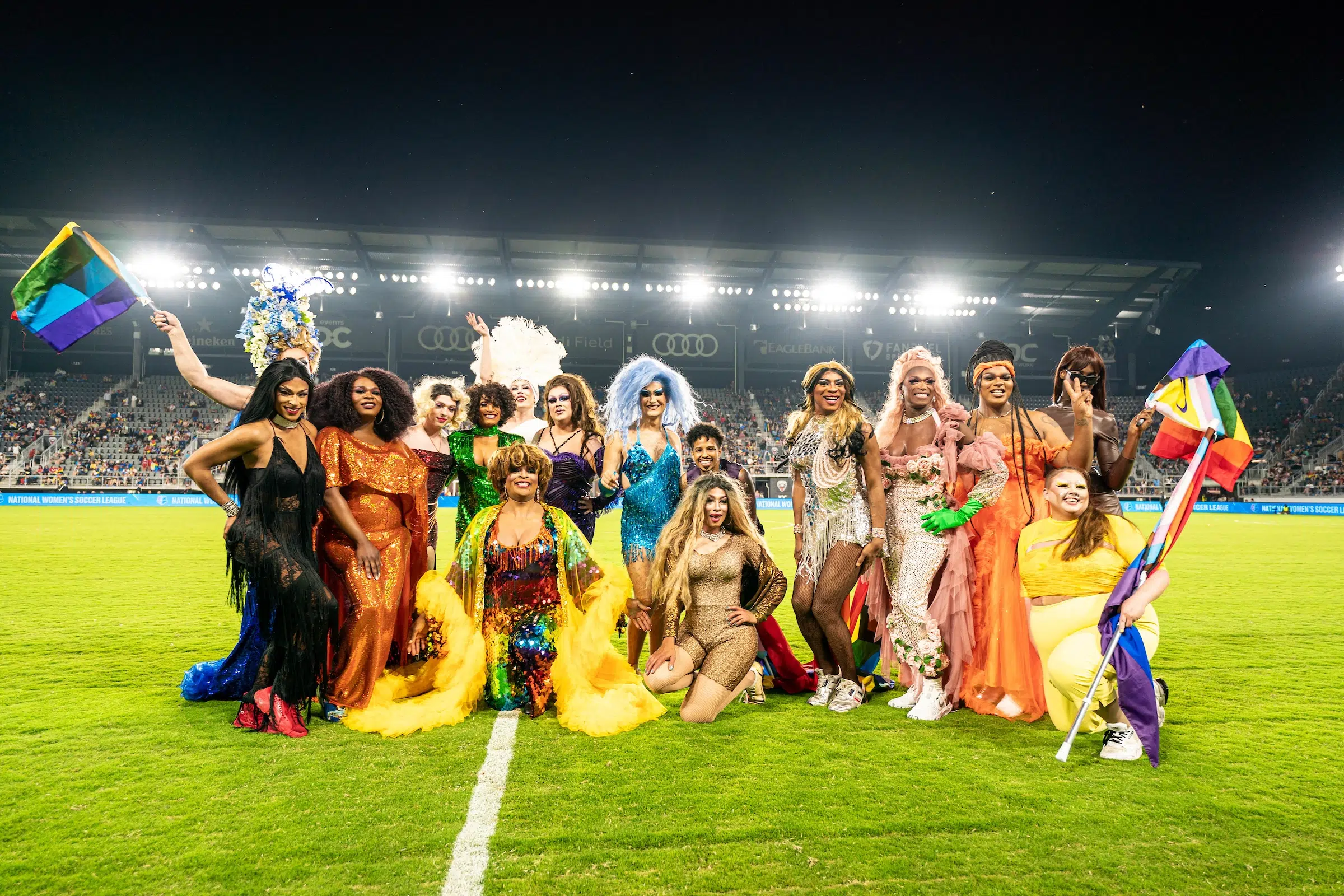 More than a dozen drag queens wearing a variety of colors, sequins, and glitter pose for photo at midfield.