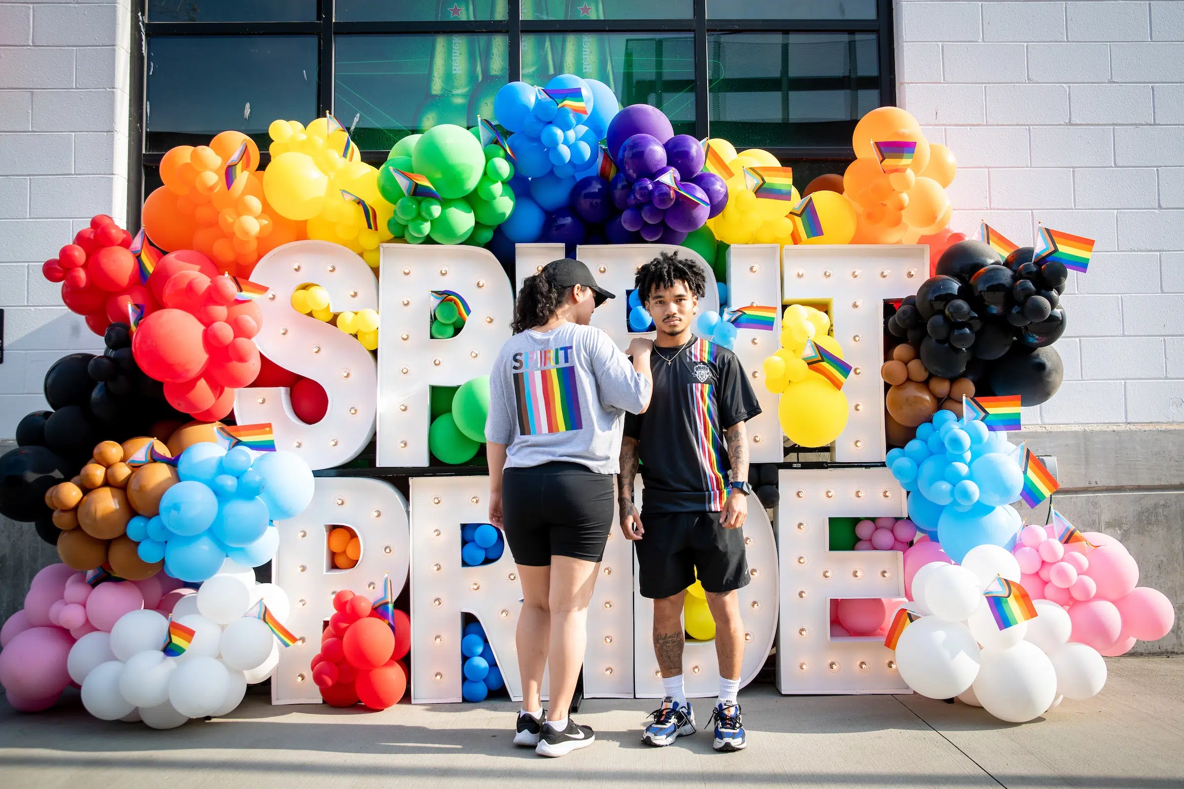 A fan in a gray "Pride" t-shirt and one in a black "Pride" t-shirt pose in front of a rainbow display of balloons and giant white, marquee letters that spell out "SPIRIT PRIDE".