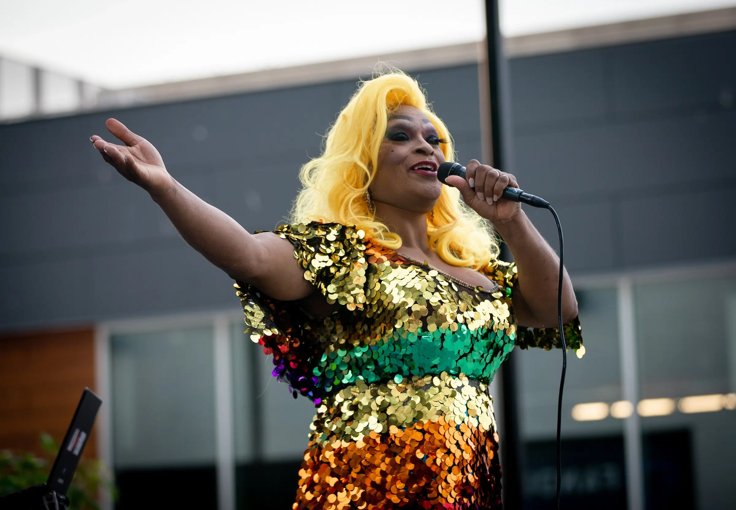 A drag queen in a rainbow sequin dress and a blonde wig speaks into a microphone.