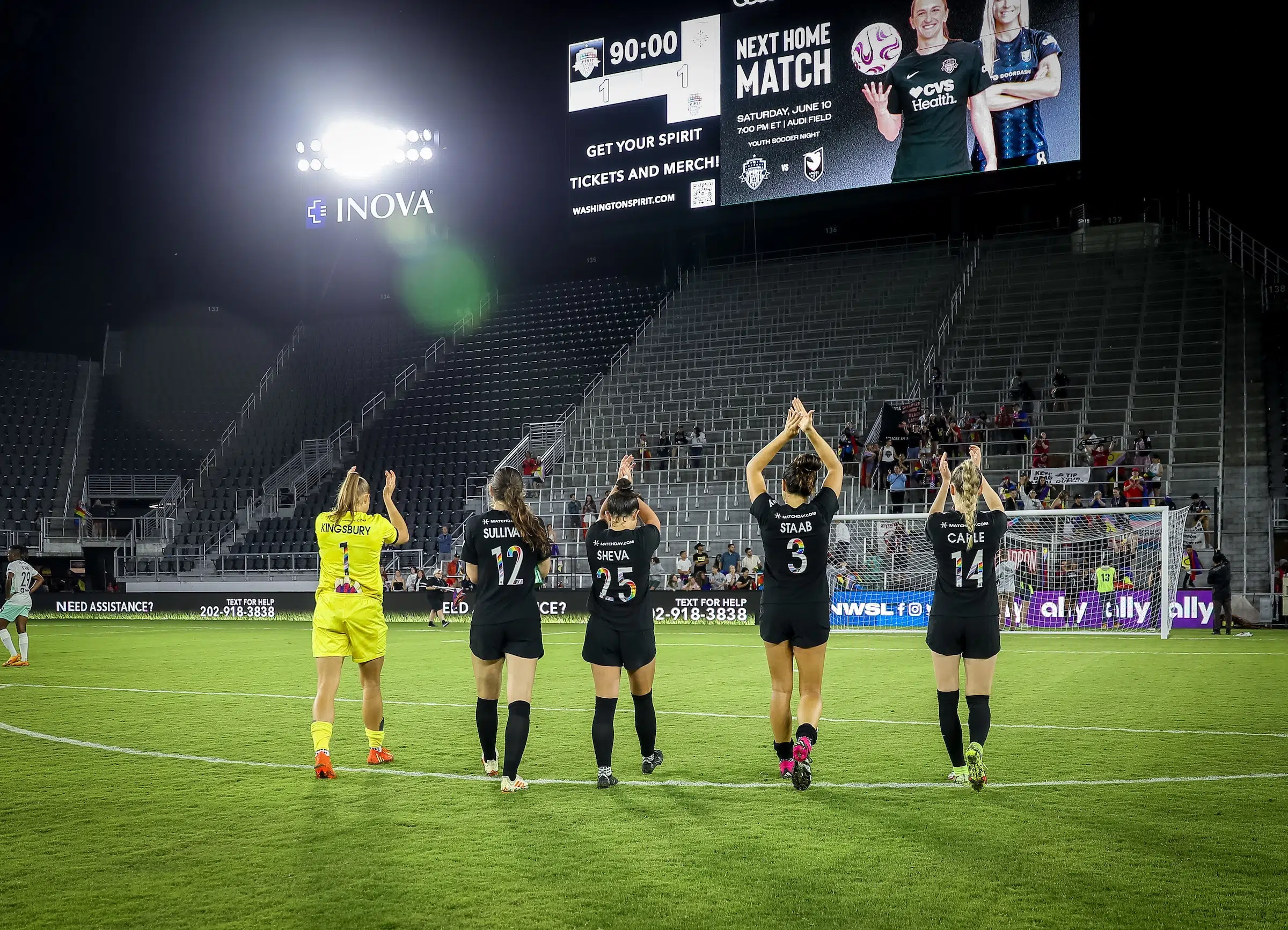 Four players in black uniforms and one in a yellow uniform clap their hands over their heads in appreciation to fans in the stands.