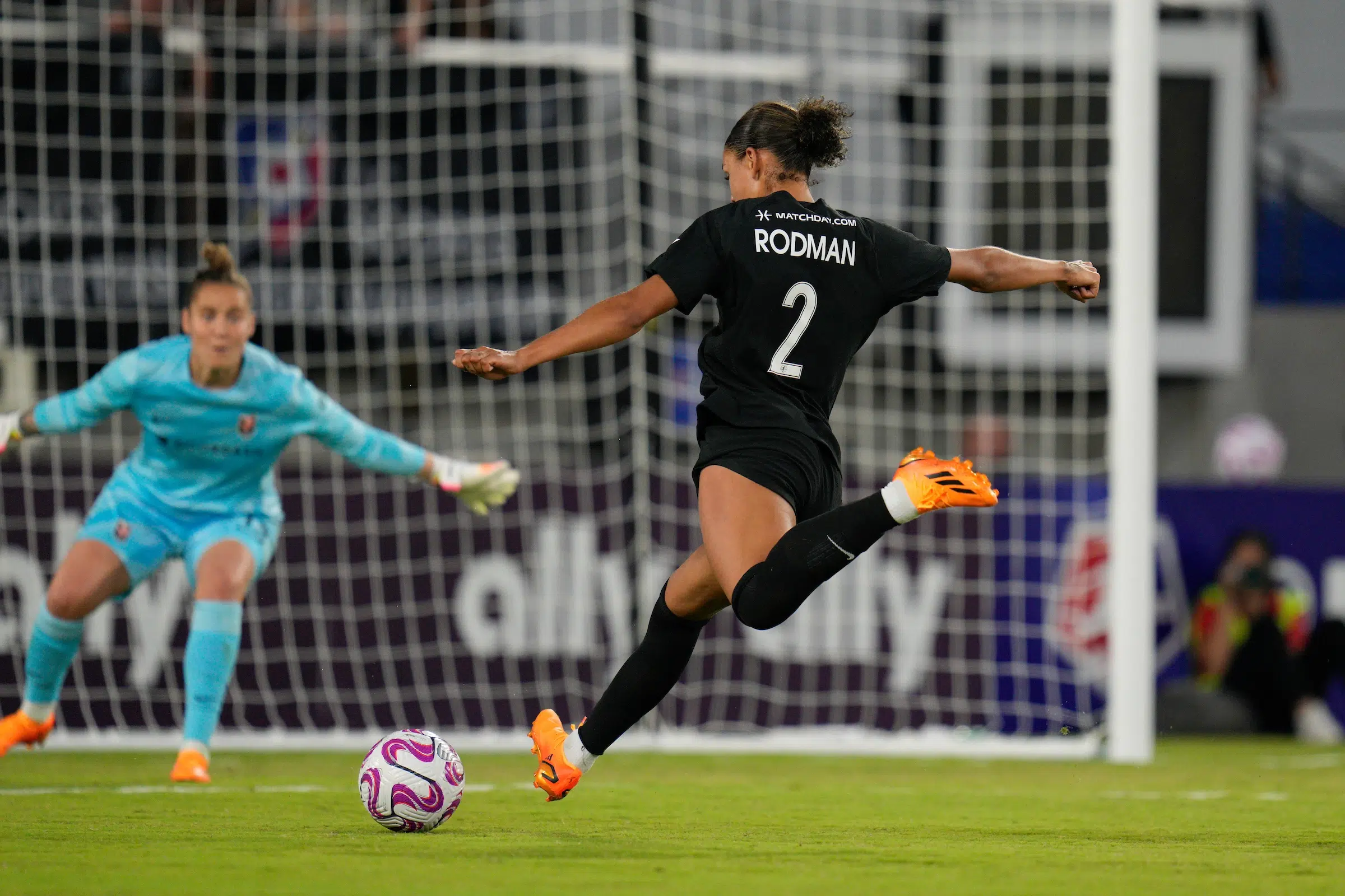 In the foreground, Trinity Rodman in an all black uniform shoots a soccer ball. In the background out of focus, a goalie in a bright blue uniform outstretches her arms and attempts to block the shot.