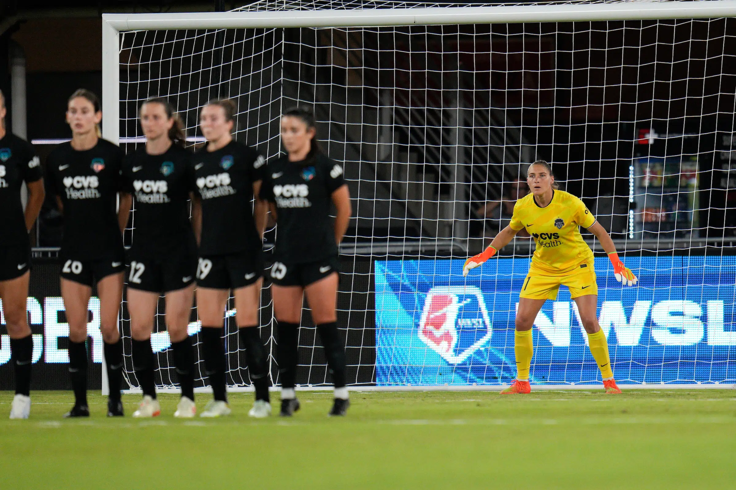 In focus in the background, Aubrey Kingsbury in a yellow uniform prepares for a free kick to be shot. In the foreground out of focus, five players in black uniforms form a wall to block the shot.