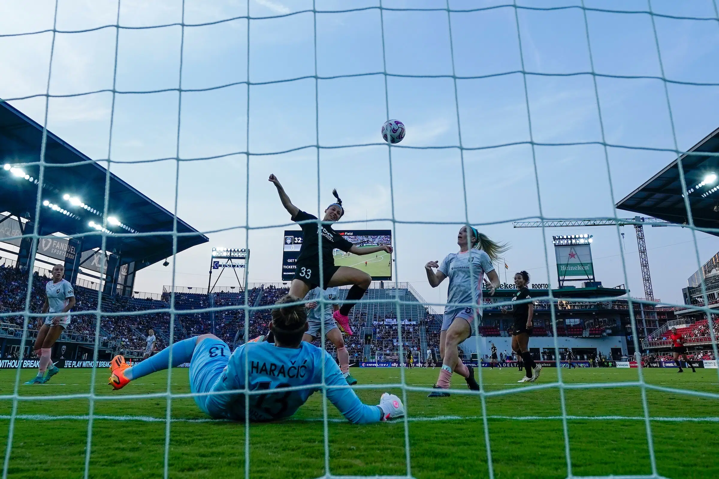 In a photo taken from behind a soccer goal, Ashley Hatch heads a soccer ball into the goal as a goalie in a blue uniform lies on the ground.