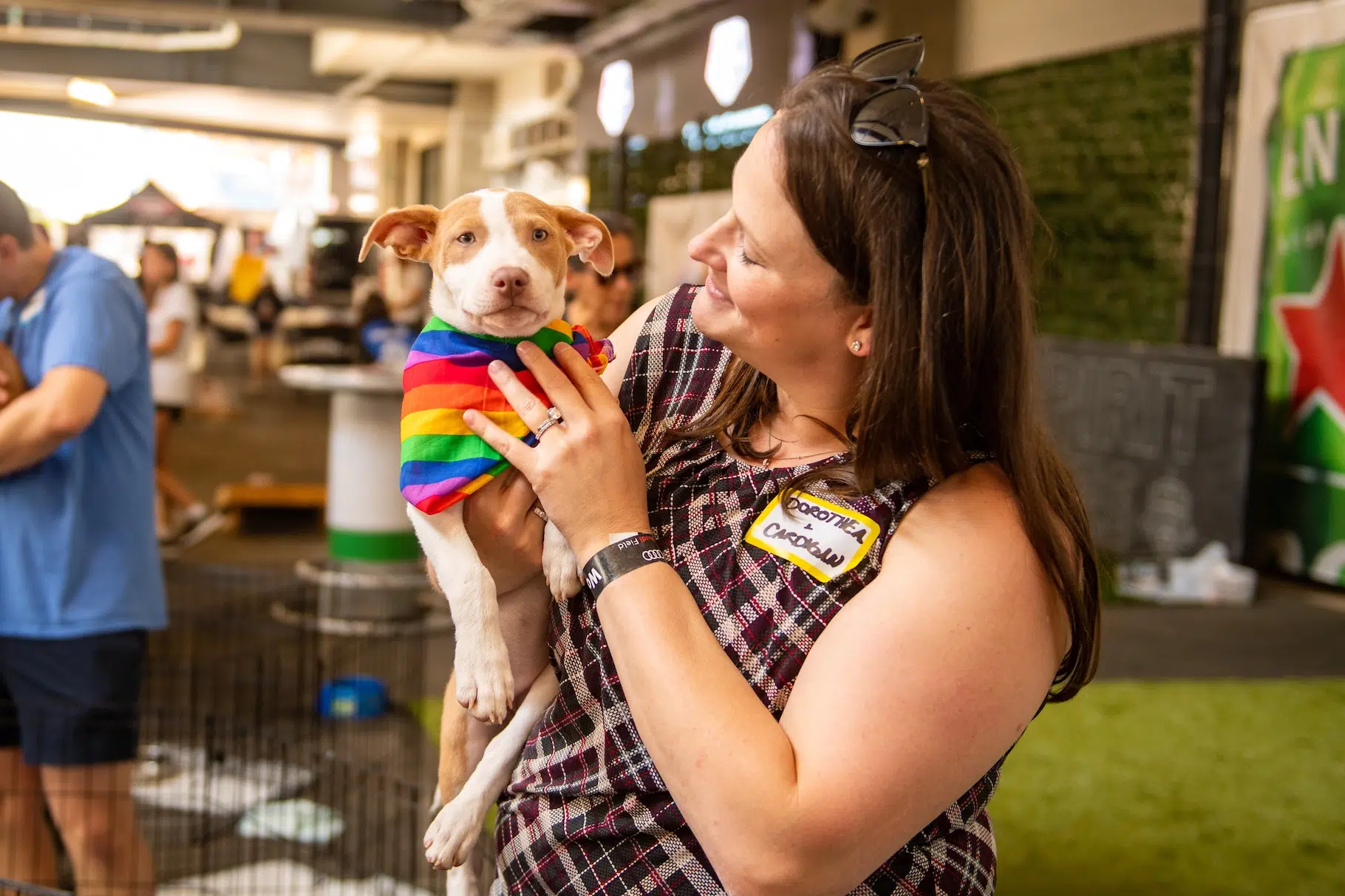 A woman in a plaid tank top holds a light brown and white dog with a rainbow bandana.